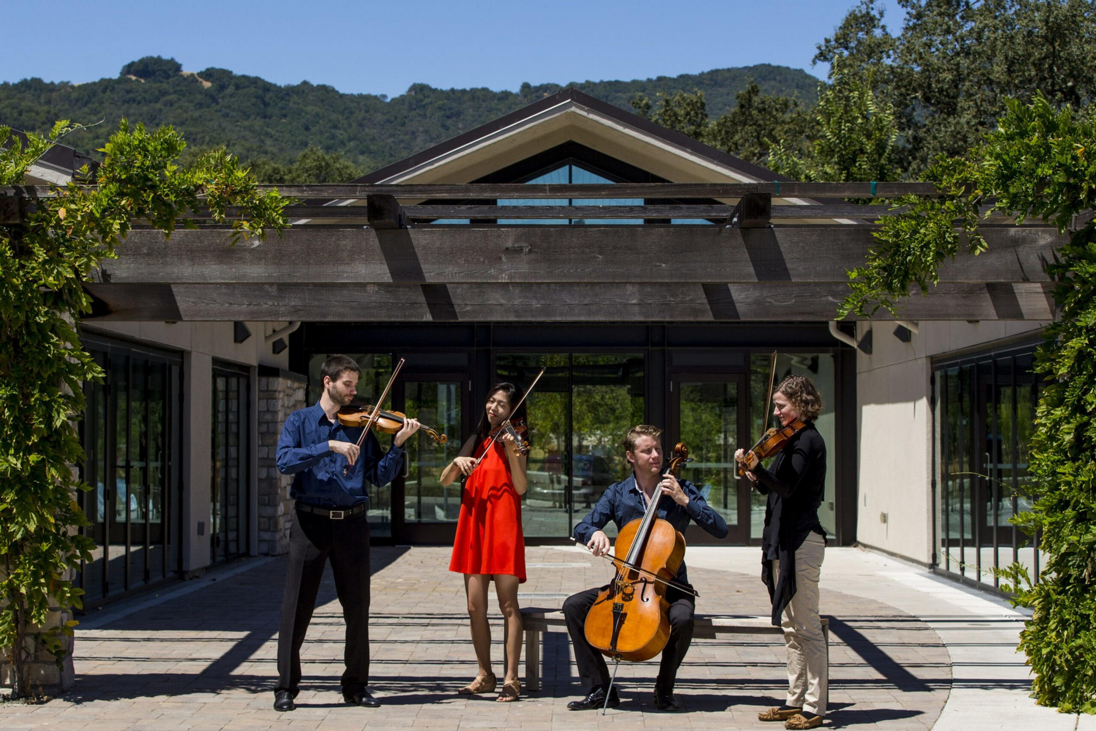 Four musicians play string instruments outside a modern building under a wooden pergola with greenery and hills in the background.