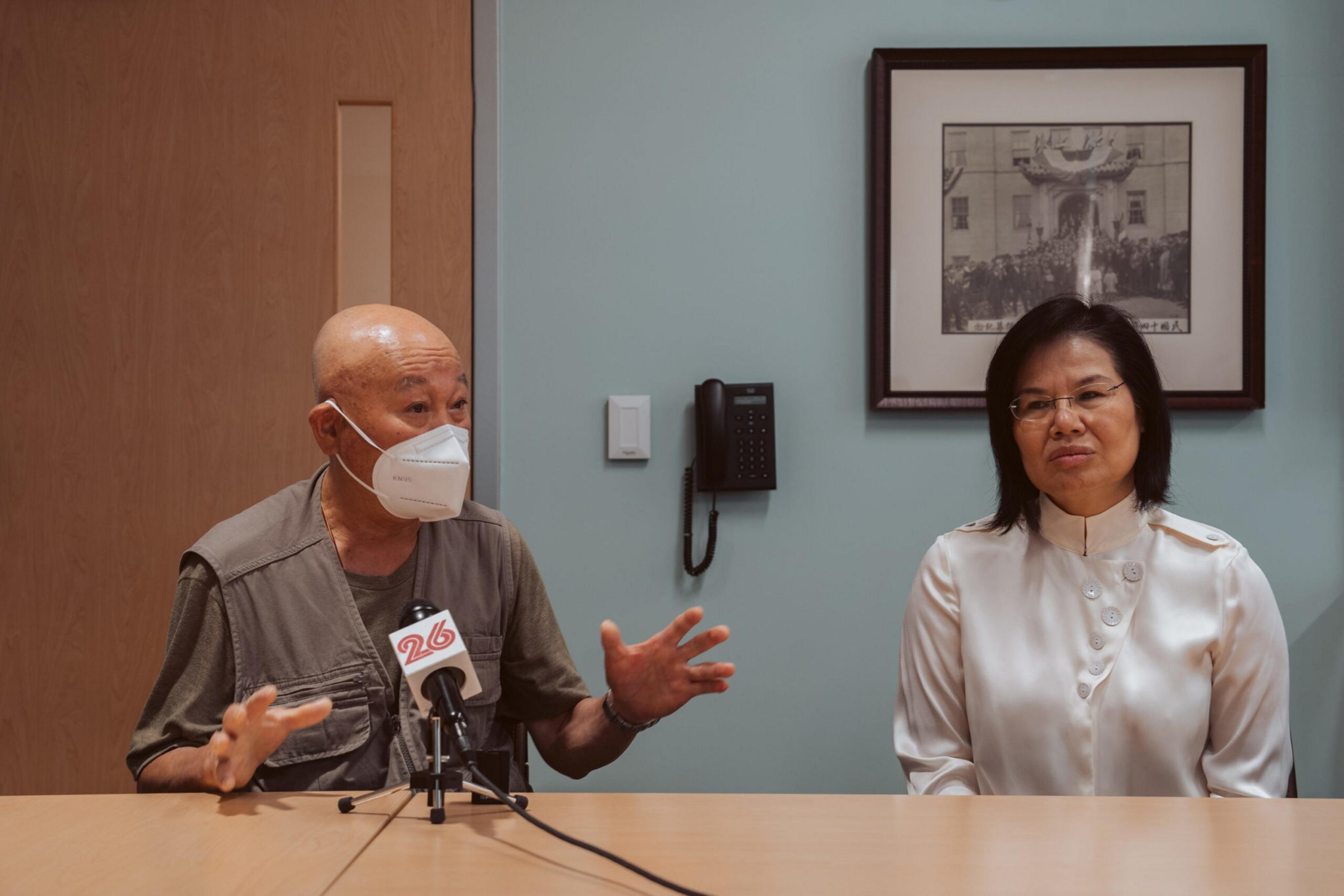 A man wearing a mask gestures while speaking into a microphone, sitting next to a woman in a white blouse, both at a table in a room with a phone and framed photo on the wall.