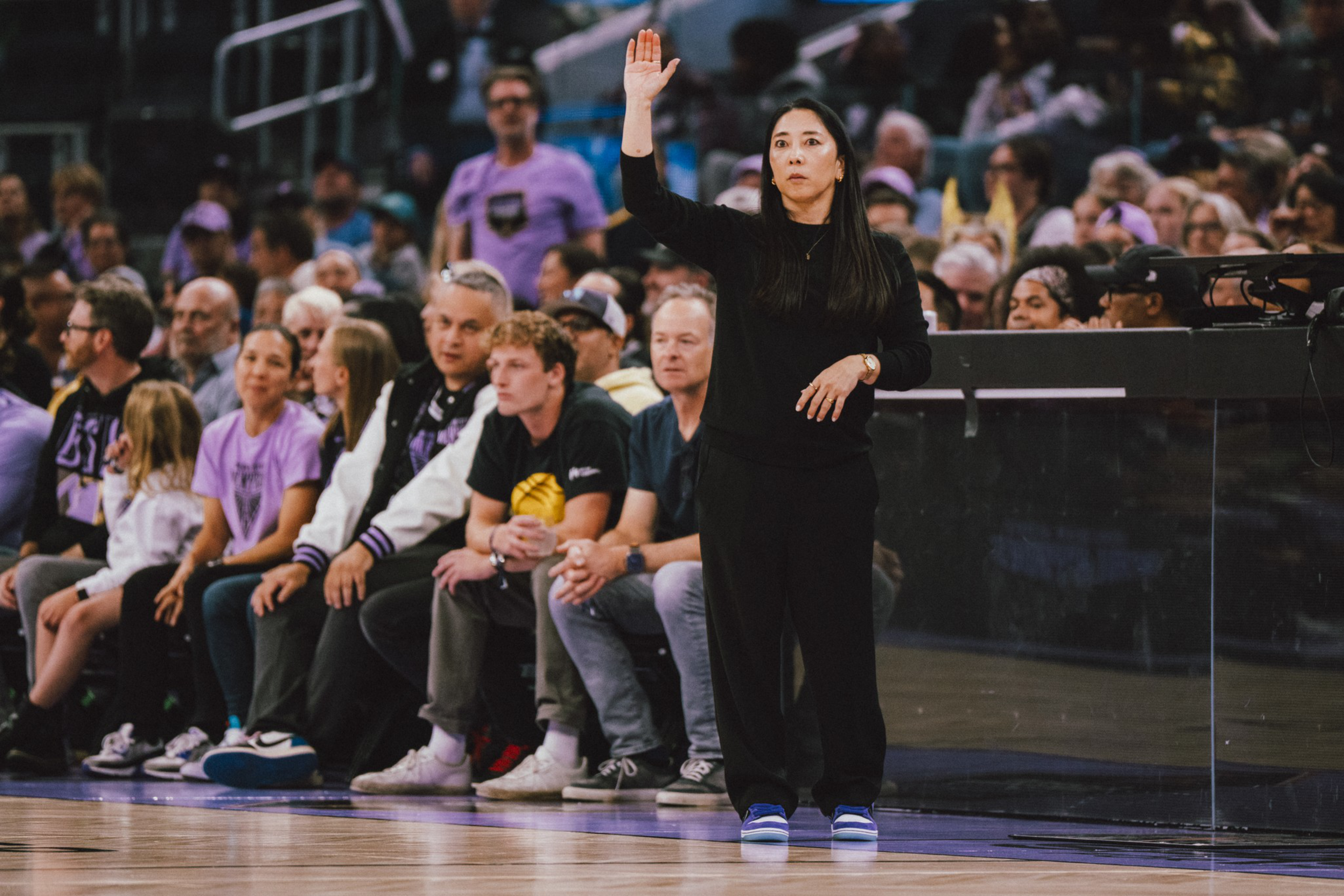 A woman dressed in black raises her right hand while standing on a basketball court, with a seated and attentive crowd in the background.