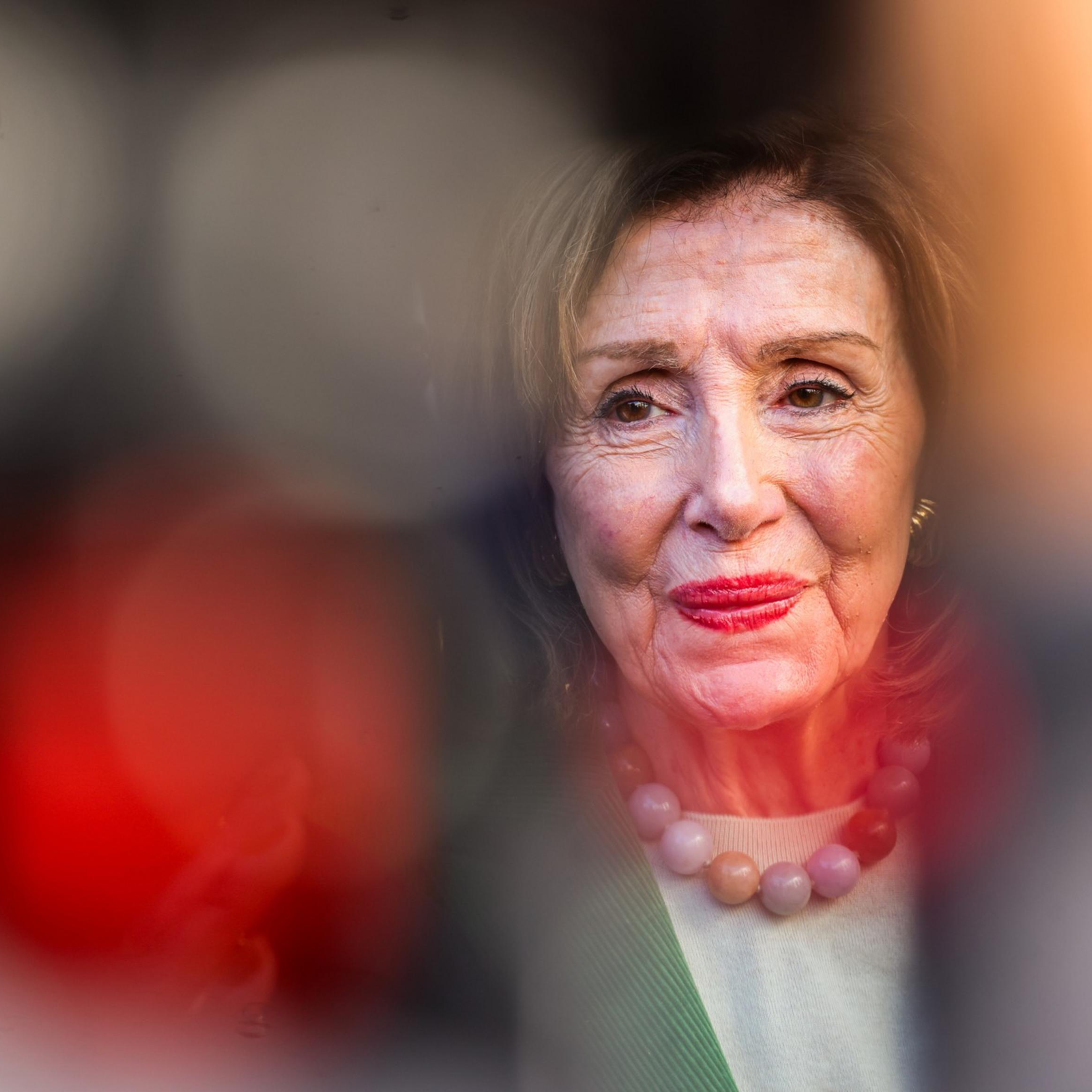 An elderly woman with light skin, wearing a pale green top and a beaded necklace, has a gentle smile and red lipstick in soft lighting.