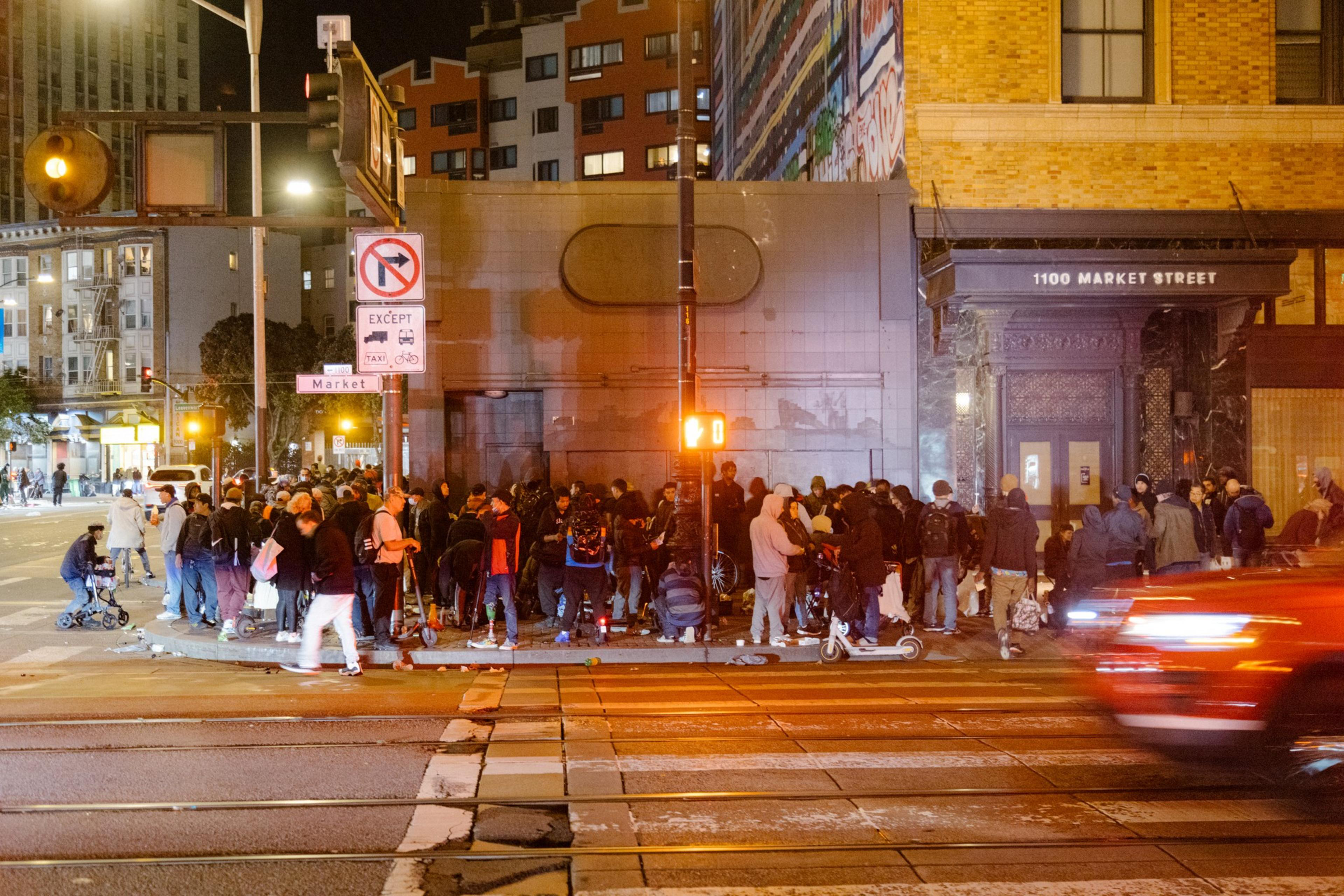A crowded city street at night with people gathered and a car passing by.