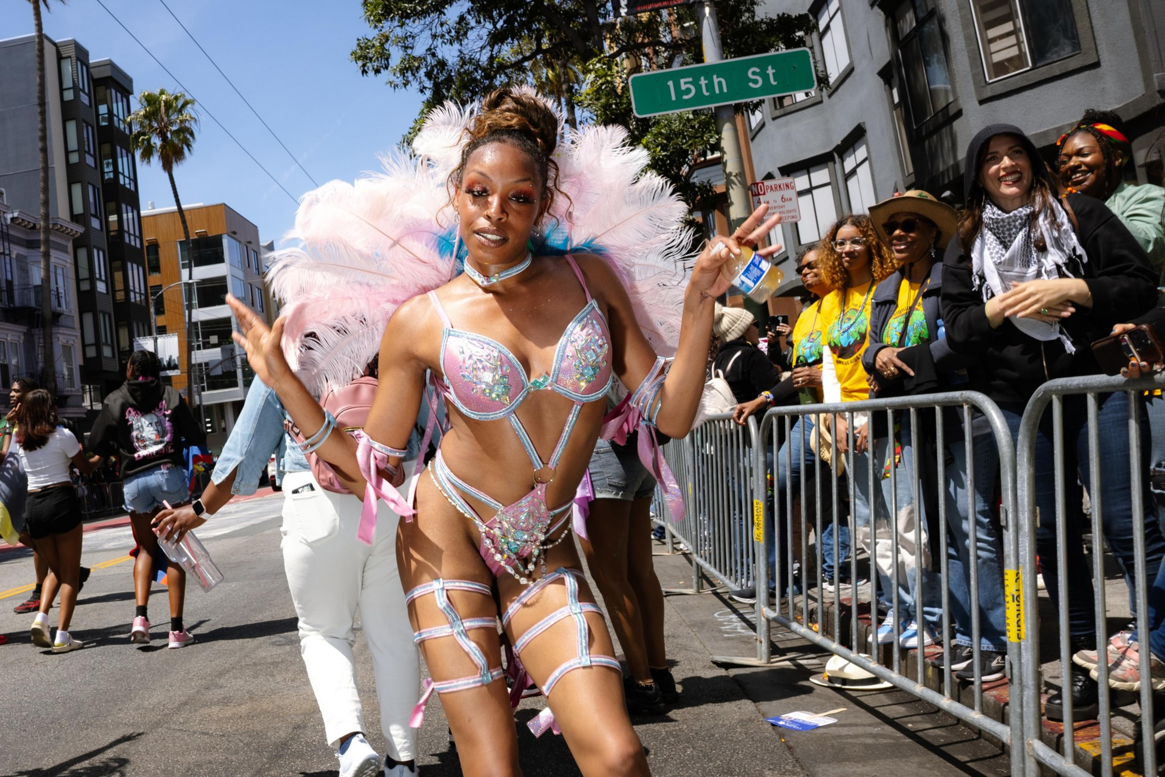 A person in a sparkling, feathered costume poses with peace signs on a street. A crowd watches from behind a barrier. Buildings and a palm tree are in the background.