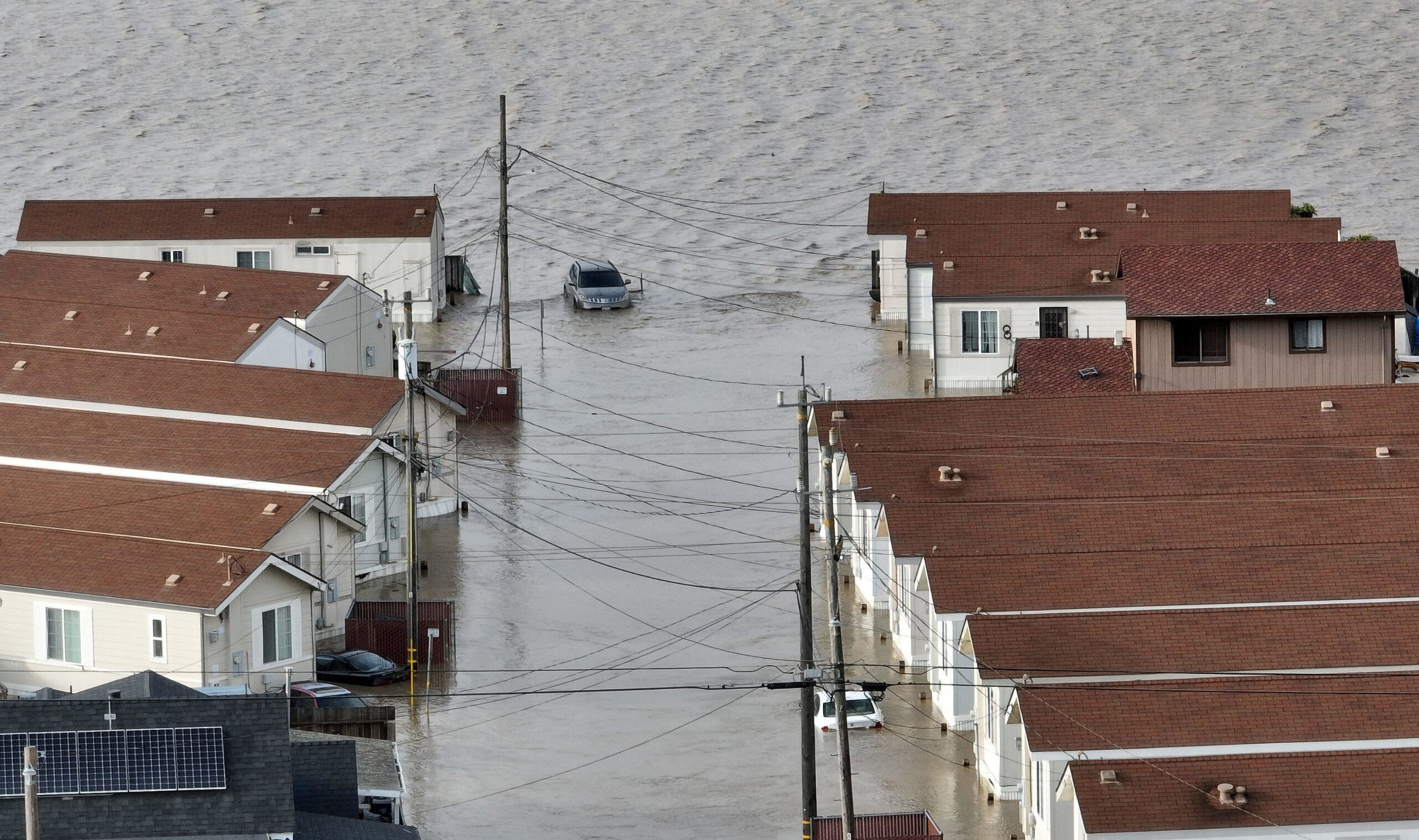 Several houses with red roofs are partially submerged in floodwater, with cars also half underwater on the flooded street between the rows of homes.