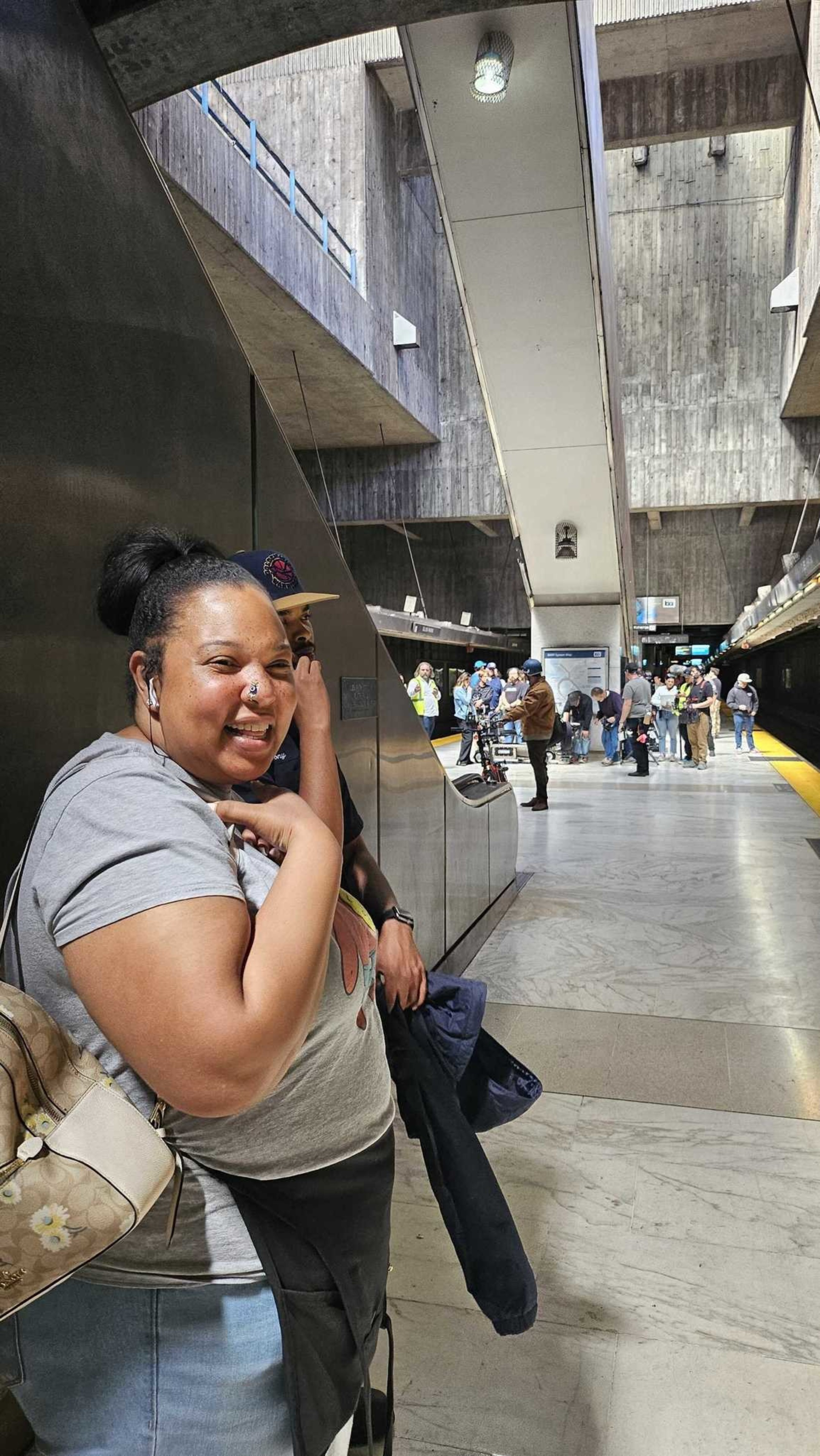 A woman smiling and wearing earbuds stands in a busy train station. Behind her, a group of people gather on the platform, which features modern concrete architecture.