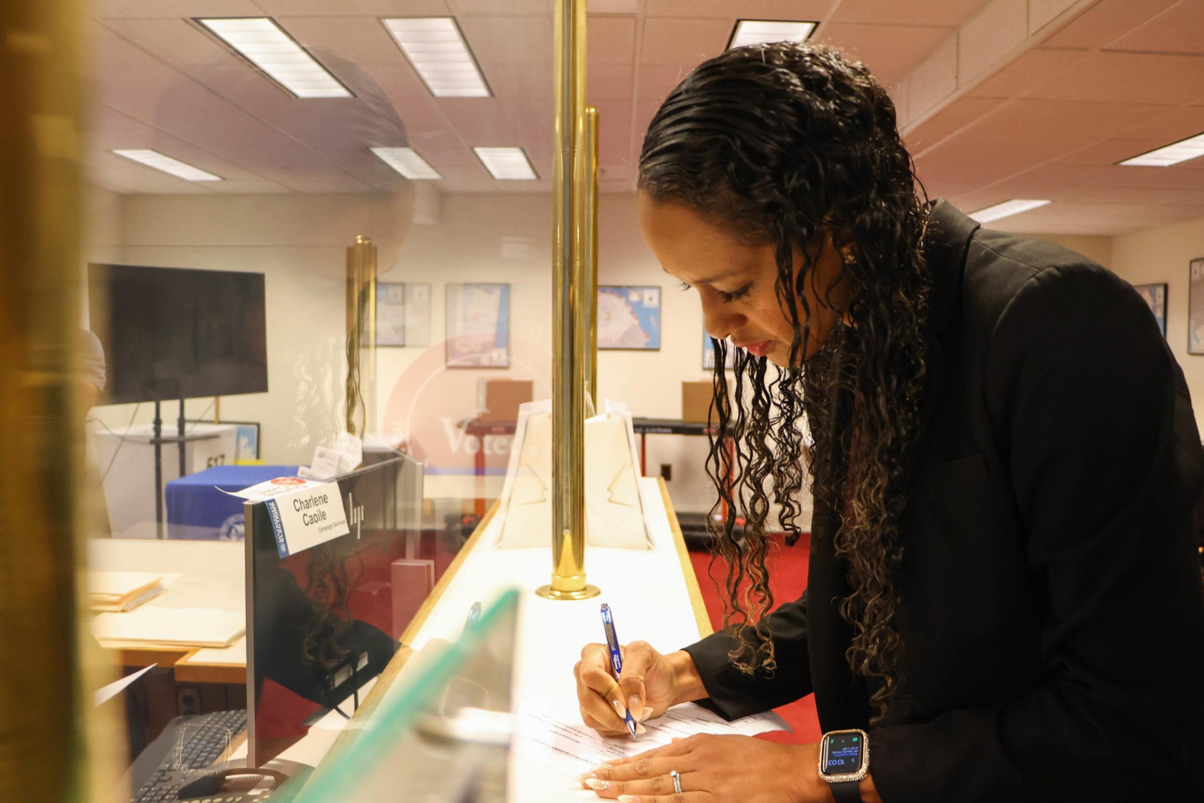 A woman with curly hair and a smartwatch is leaning over a counter, signing documents behind a glass partition in an office setting.
