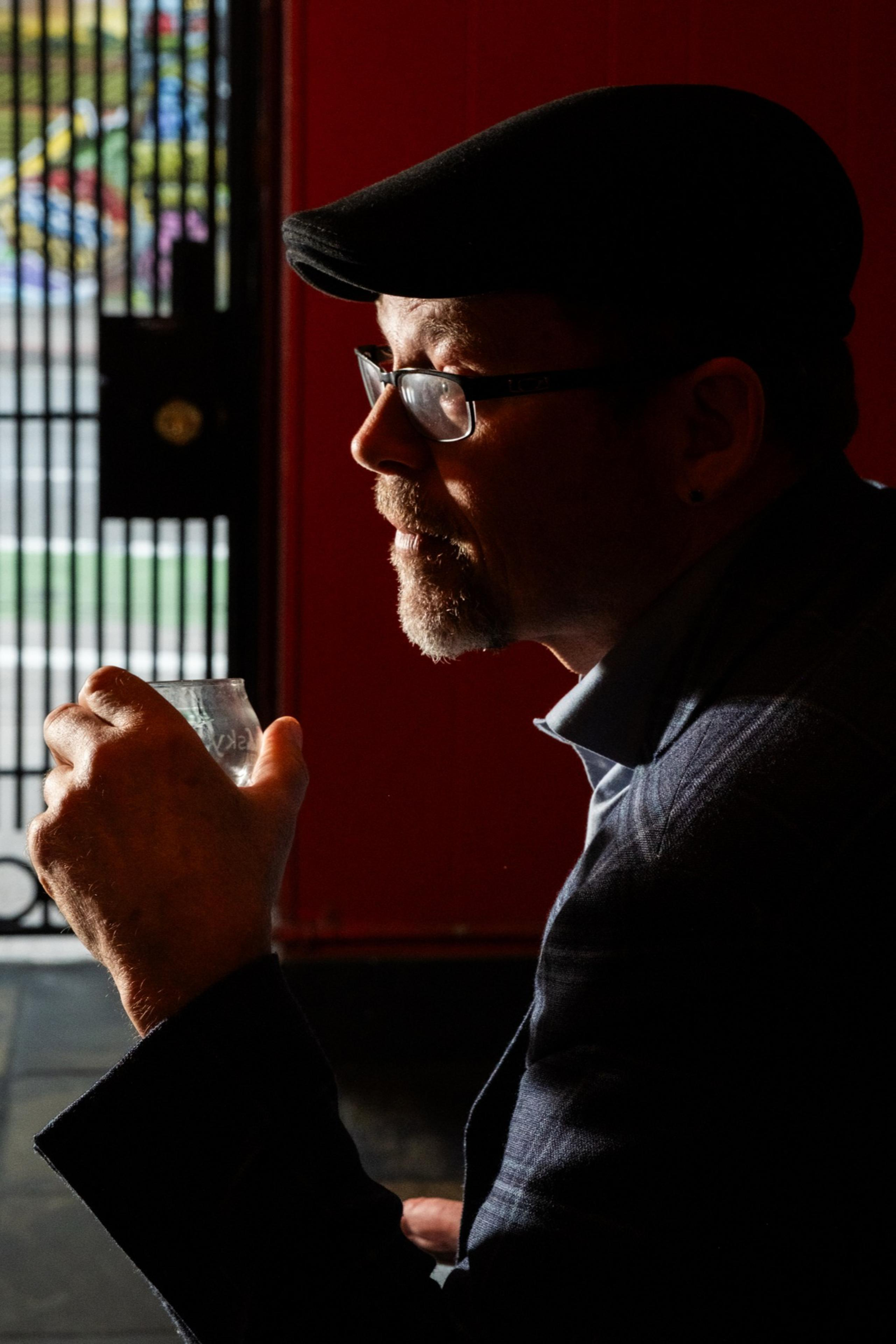 A man in a cap and glasses sits in profile holding a small glass. He is in a dimly lit room with a barred door and colorful background visible.