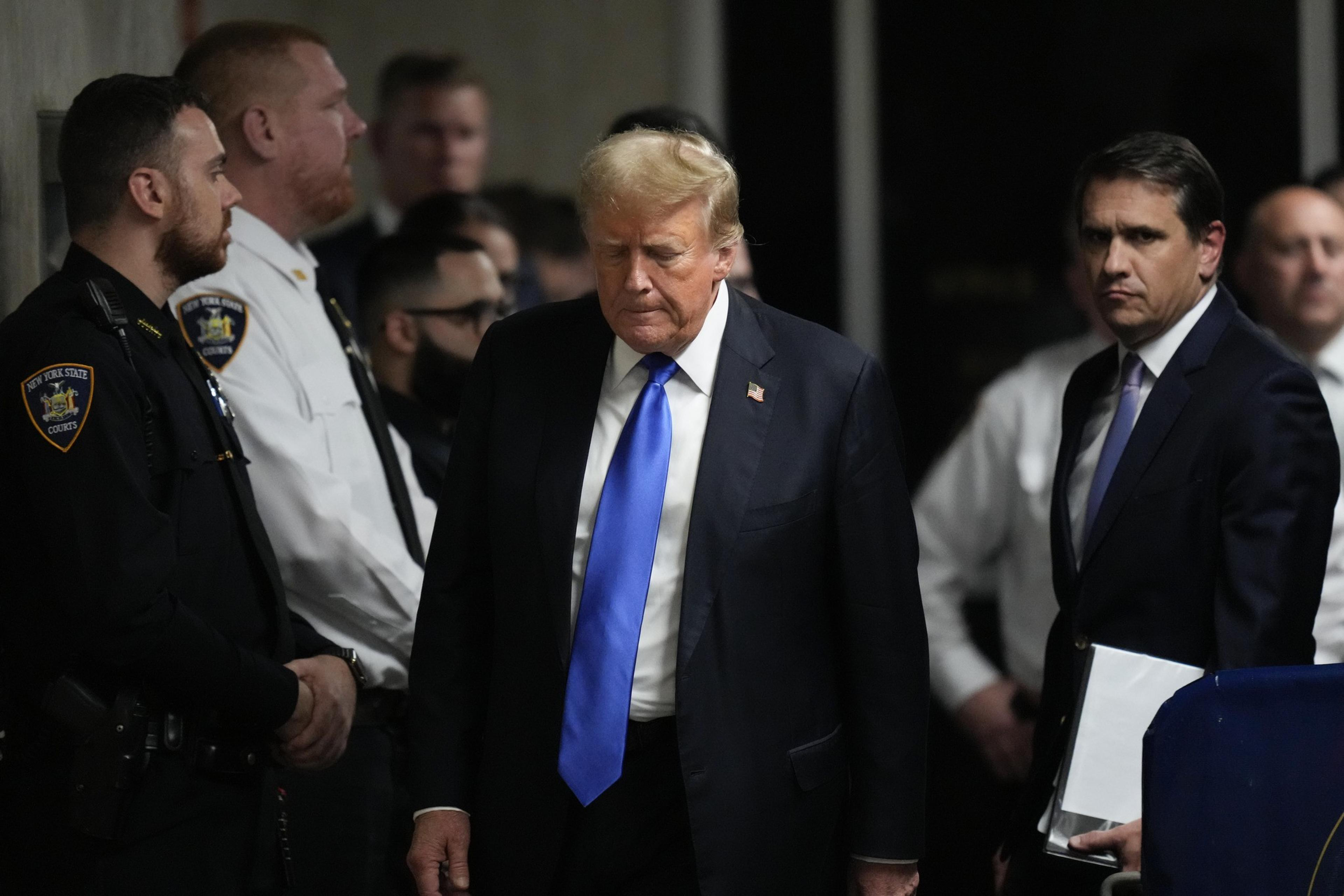 A man with blonde hair and a blue tie, flanked by security personnel and officials, walks through a corridor looking serious. He is the central figure in the image.