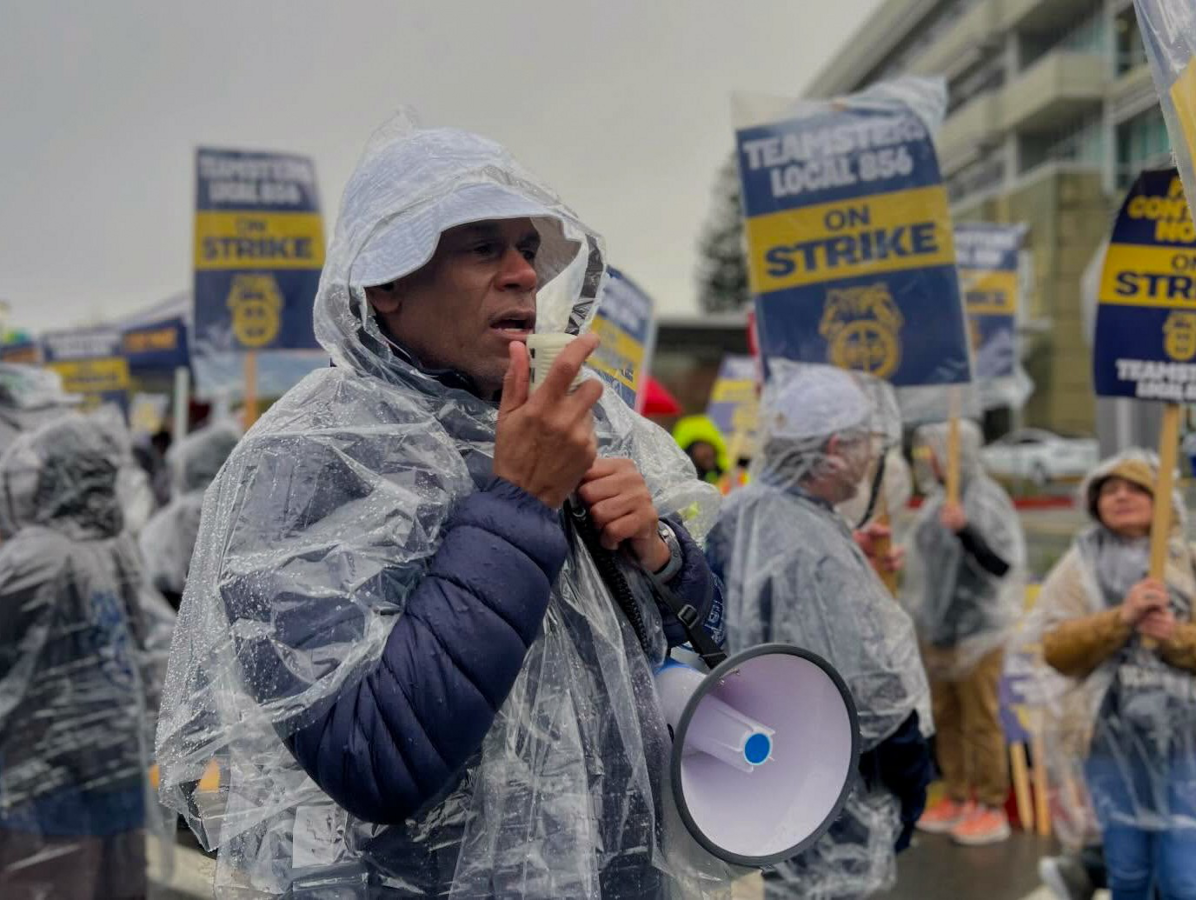 A person in a rain poncho holds a megaphone and speaks during a protest where others hold signs that say "TEAMSTERS LOCAL 856 ON STRIKE."