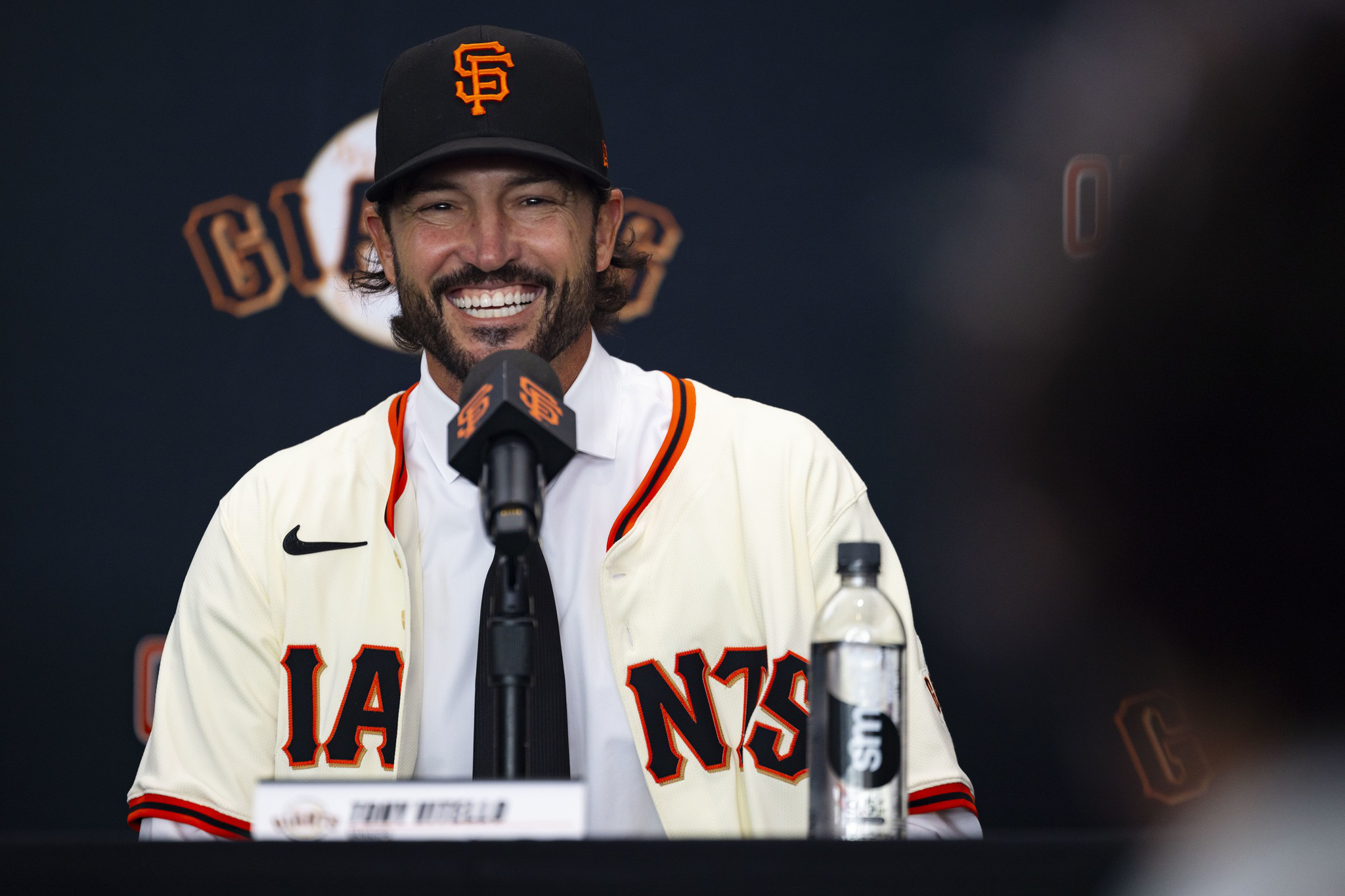 A smiling man in a San Francisco Giants jersey and cap speaks into a microphone at a press conference table with a water bottle nearby.