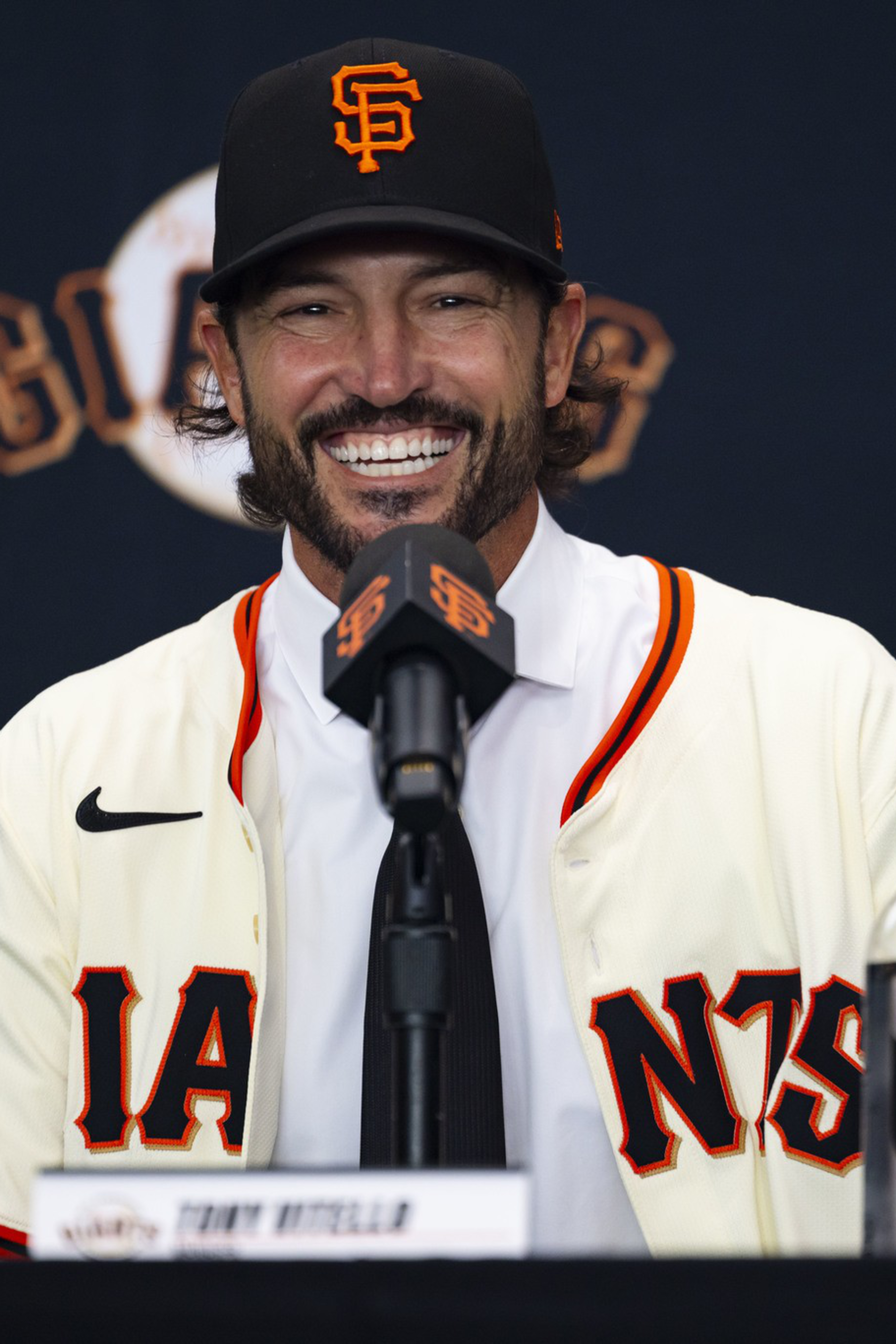 A smiling man in a San Francisco Giants jersey and cap speaks into a microphone at a press conference table with a water bottle nearby.
