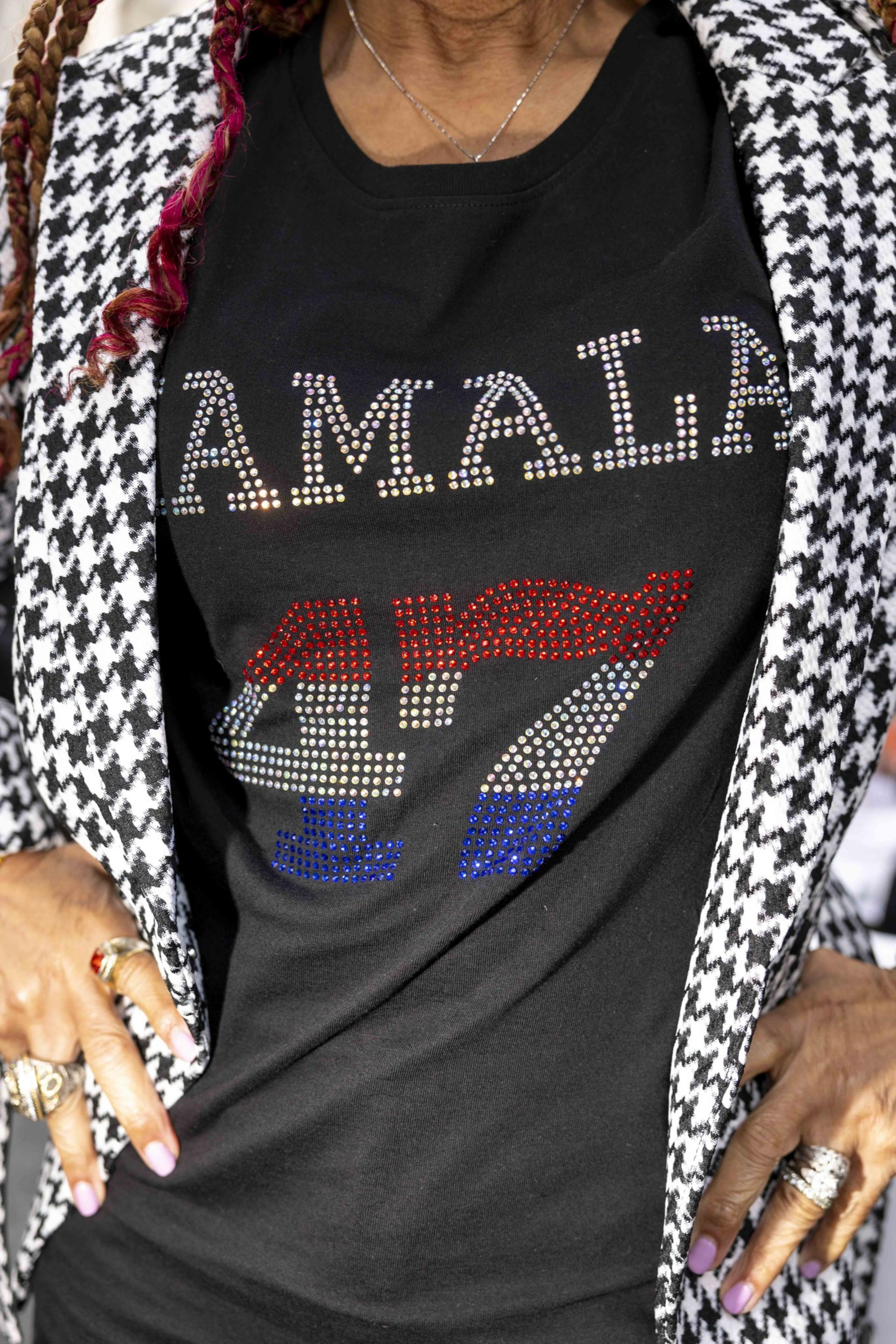 A person wears a black shirt with &quot;AMALA 47&quot; in rhinestones, under a black-and-white houndstooth jacket. They have pink nails and multiple rings.