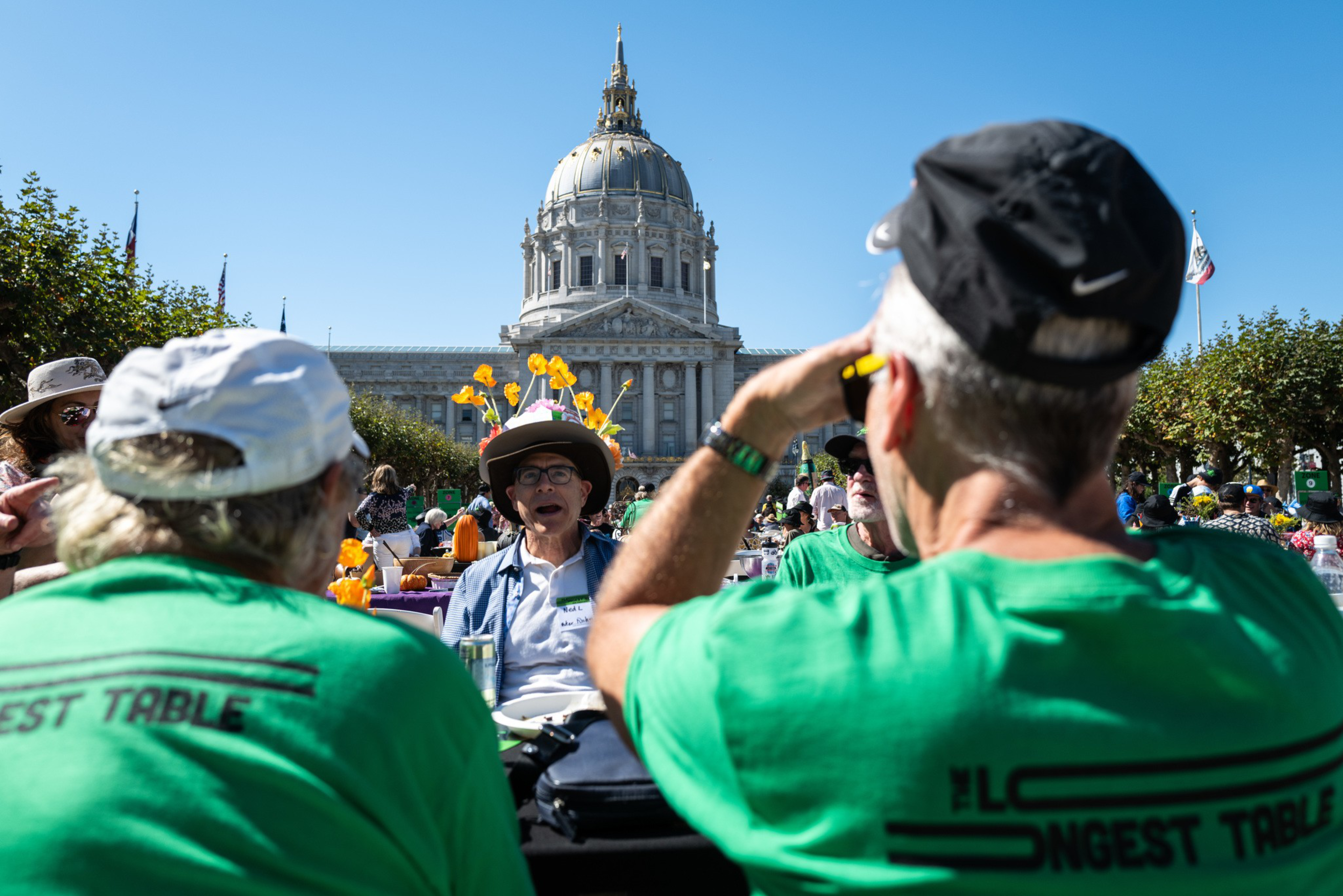 People wearing green shirts sit at an outdoor table decorated with flowers, with a large domed government building in the background under a clear blue sky.