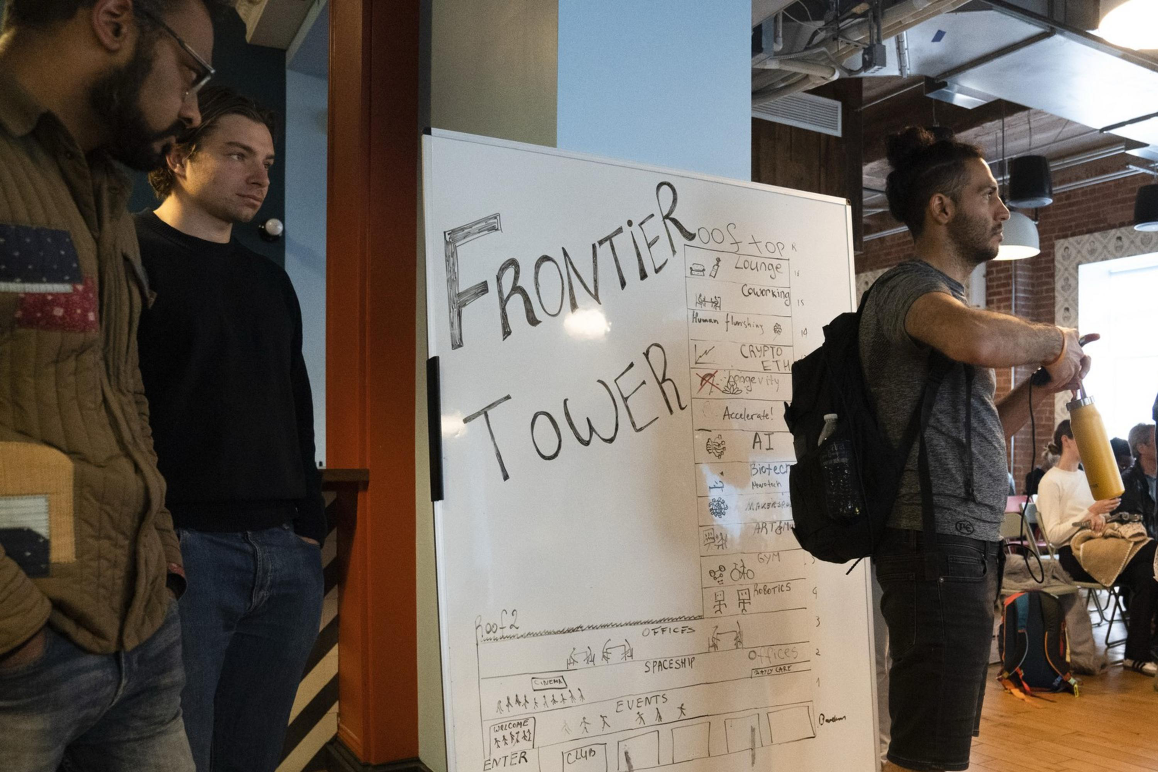 A group of people stand near a whiteboard with "FRONTIER TOWER" written on it. The room is filled with seated attendees and has a wooden floor and brick walls.