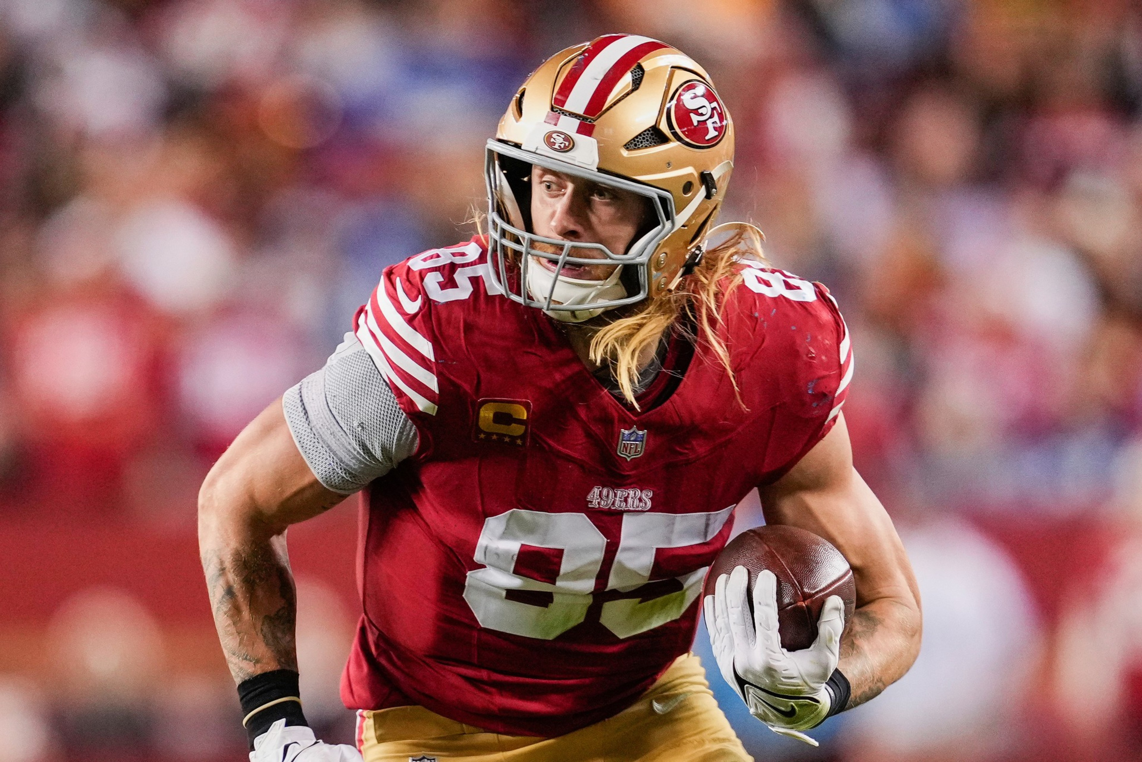 A football player in a red and gold uniform, number 85, is running with the ball. He has long blond hair and is on a blurred field with a crowd.
