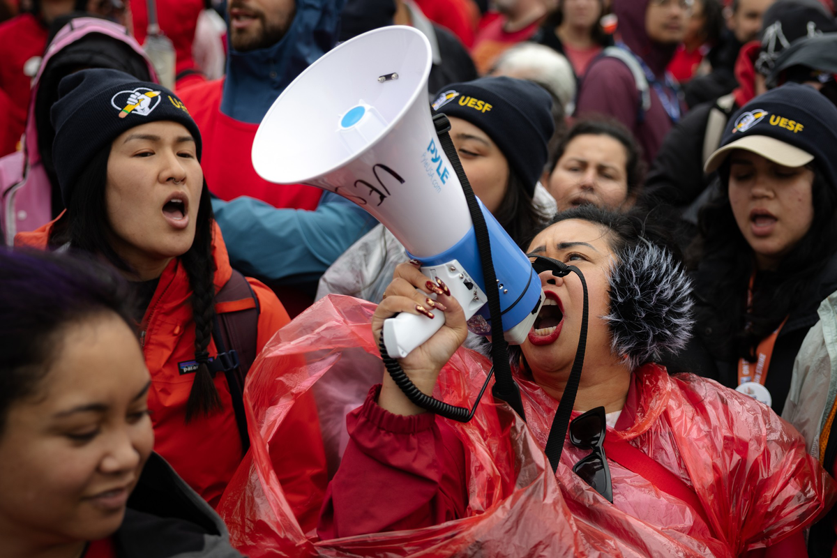 A person in a red poncho and black earmuffs shouts into a megaphone while surrounded by people, some wearing black beanies with UESF logos.