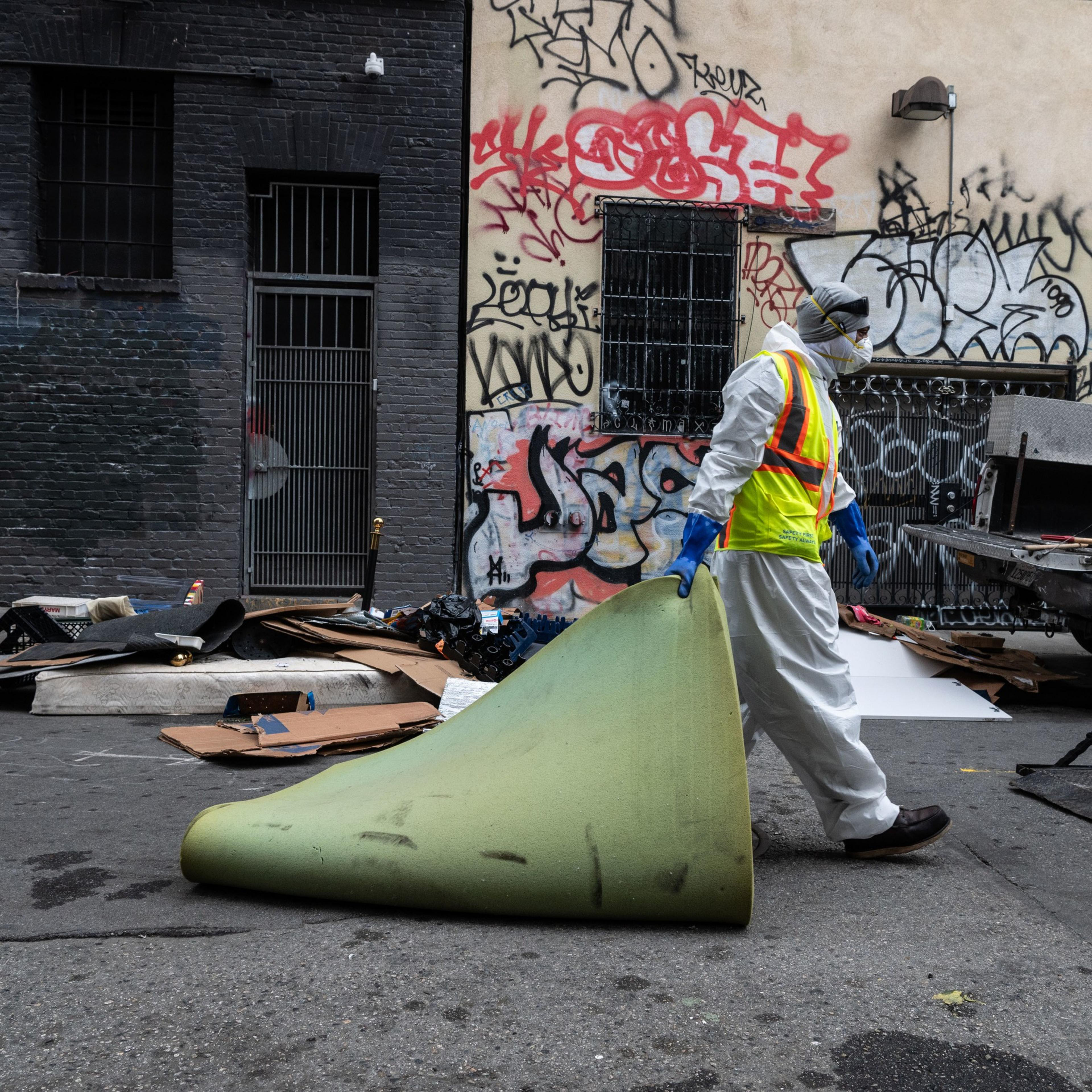 A person in protective gear and a reflective vest carries a large green object in an alley filled with debris. The walls are covered in colorful graffiti.