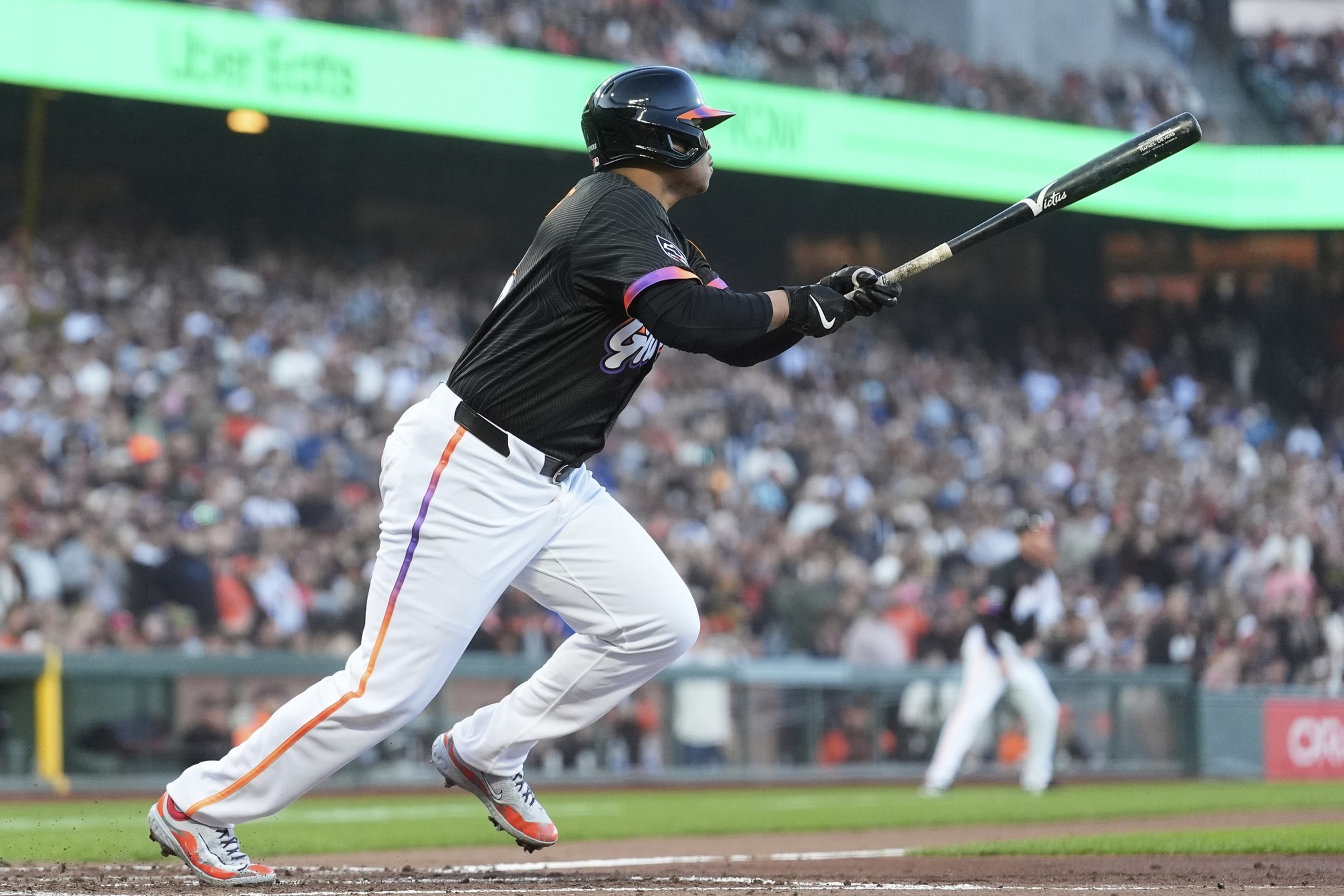 A baseball player in a black jersey and white pants is mid-swing, intently watching the ball. The crowd and stadium signage are visible in the background.