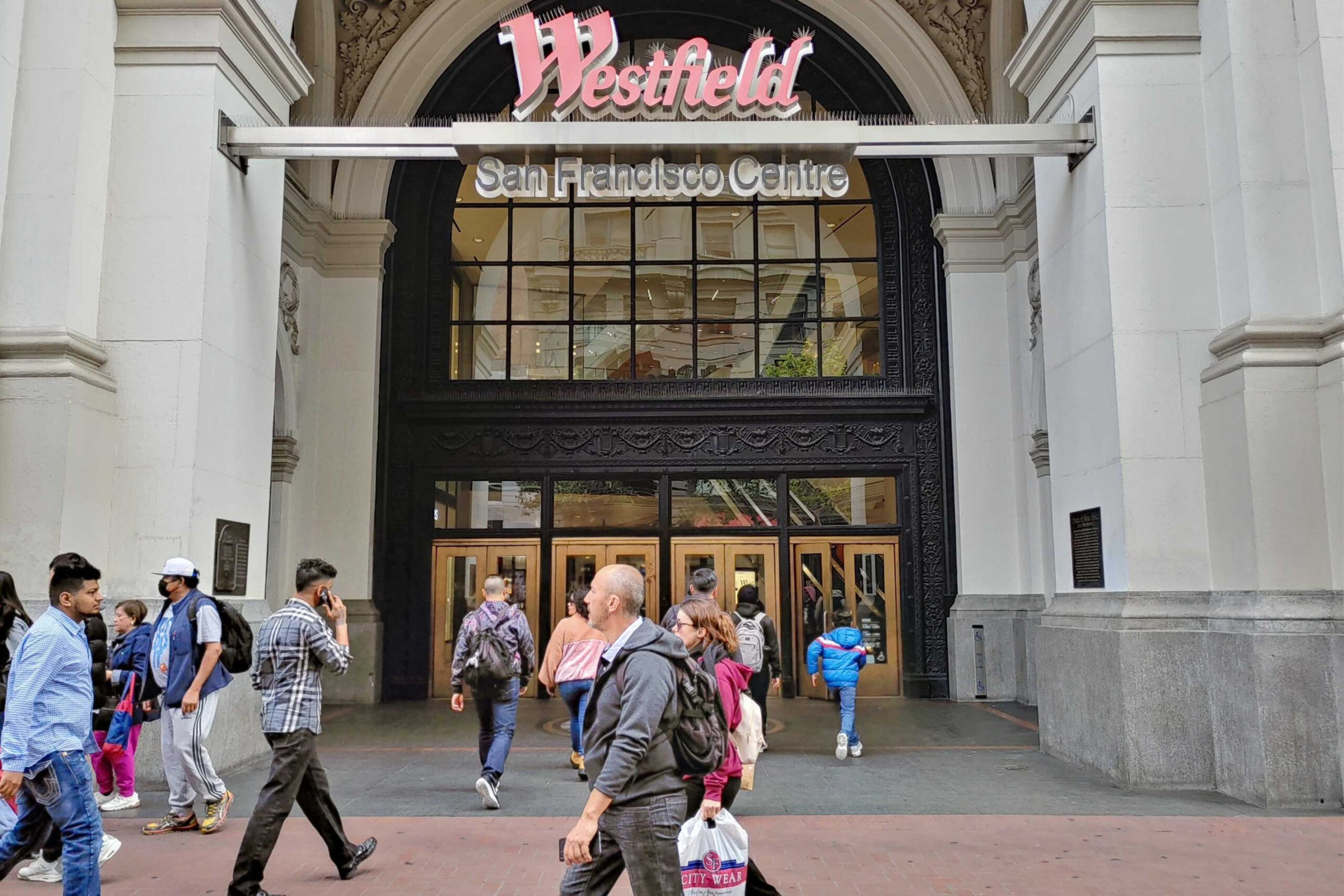 People walk in and out of the Westfield San Francisco Centre, a large shopping mall with a prominent arched entrance and multiple glass doors.