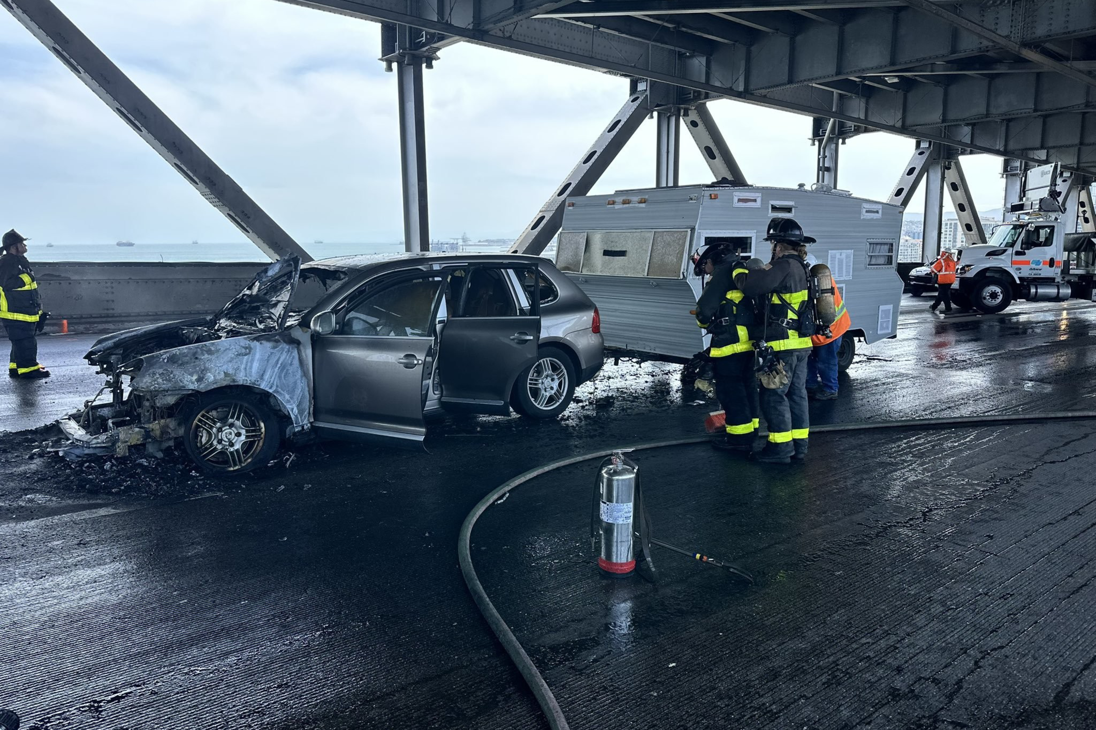 A damaged car with an open door sits on a bridge, next to a crumpled RV. Firefighters in gear inspect the scene, and an extinguisher is on the ground nearby.