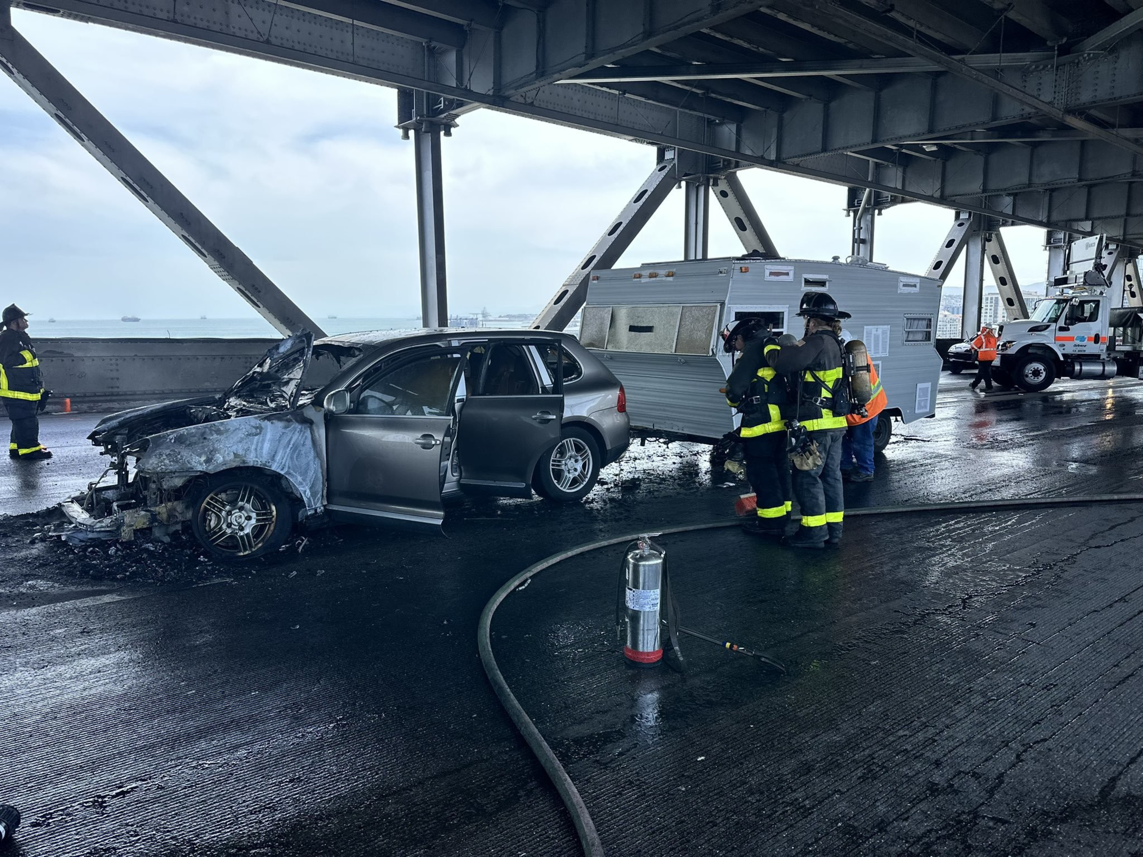 A damaged car with an open door sits on a bridge, next to a crumpled RV. Firefighters in gear inspect the scene, and an extinguisher is on the ground nearby.