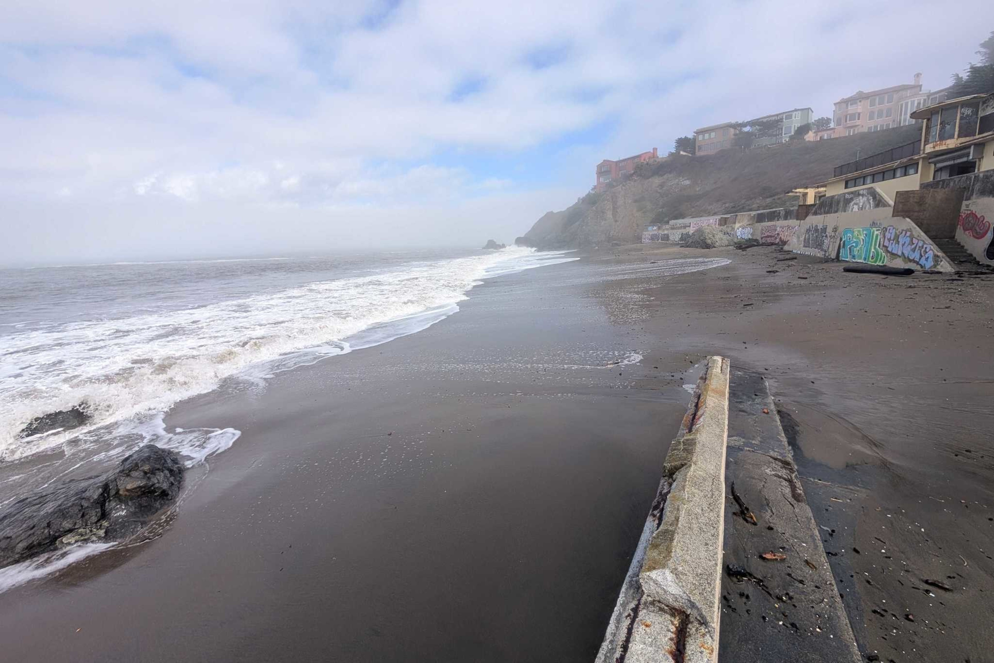 A foggy beach with waves washing ashore, rocky cliffs in the distance, and a seawall covered in graffiti lining the sandy shore.