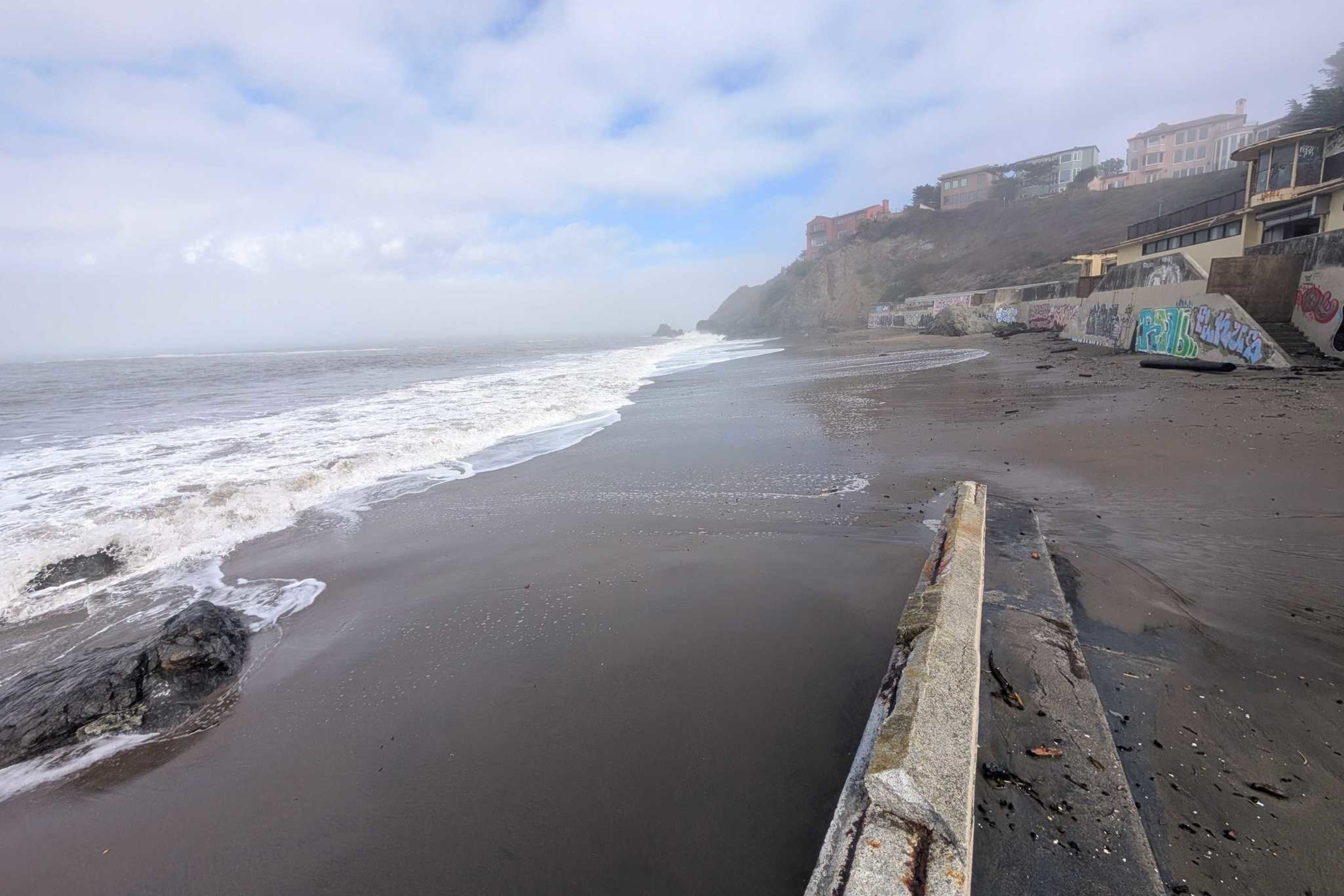 A foggy beach with waves washing ashore, rocky cliffs in the distance, and a seawall covered in graffiti lining the sandy shore.