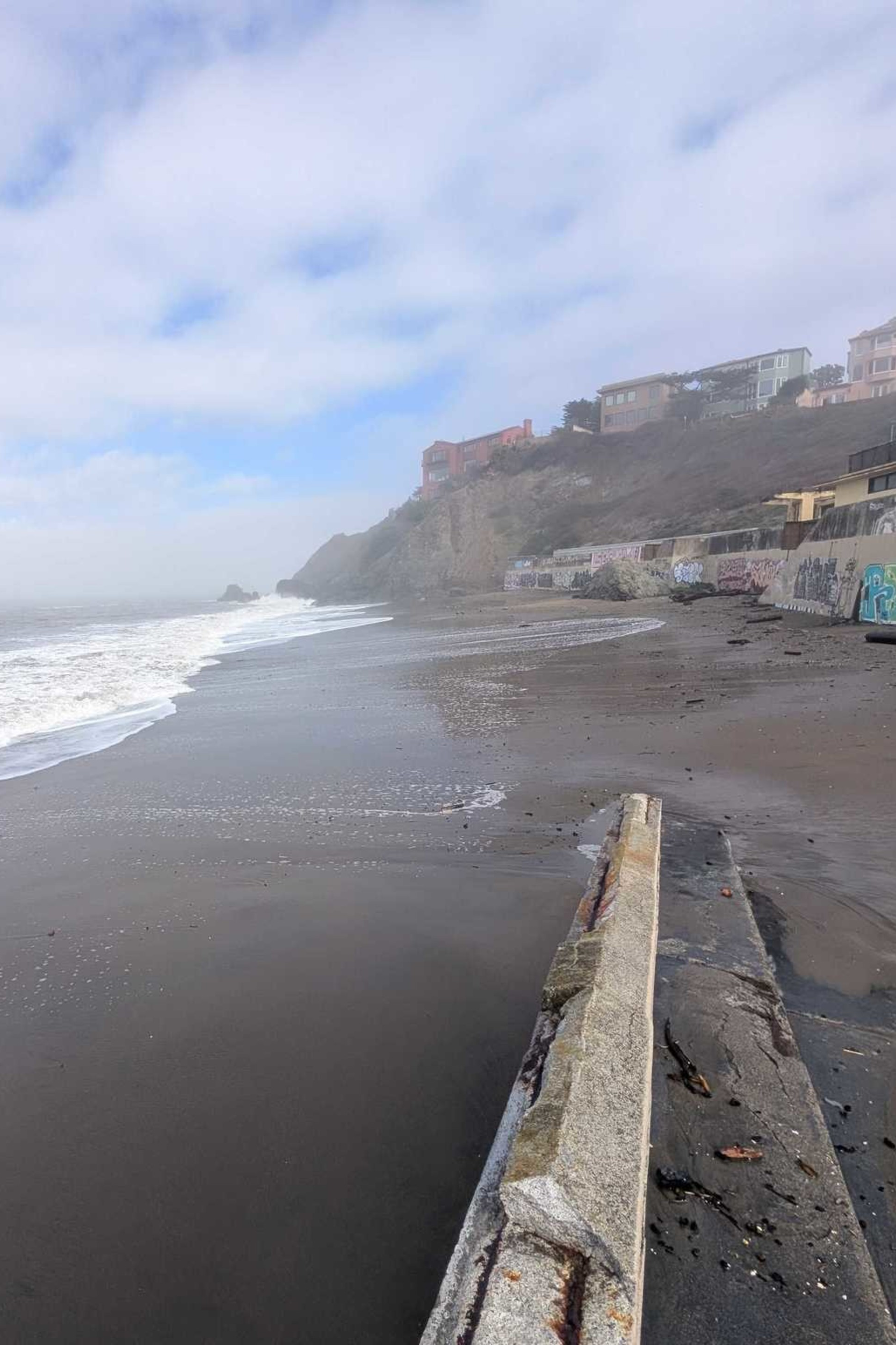 A foggy beach with waves washing ashore, rocky cliffs in the distance, and a seawall covered in graffiti lining the sandy shore.
