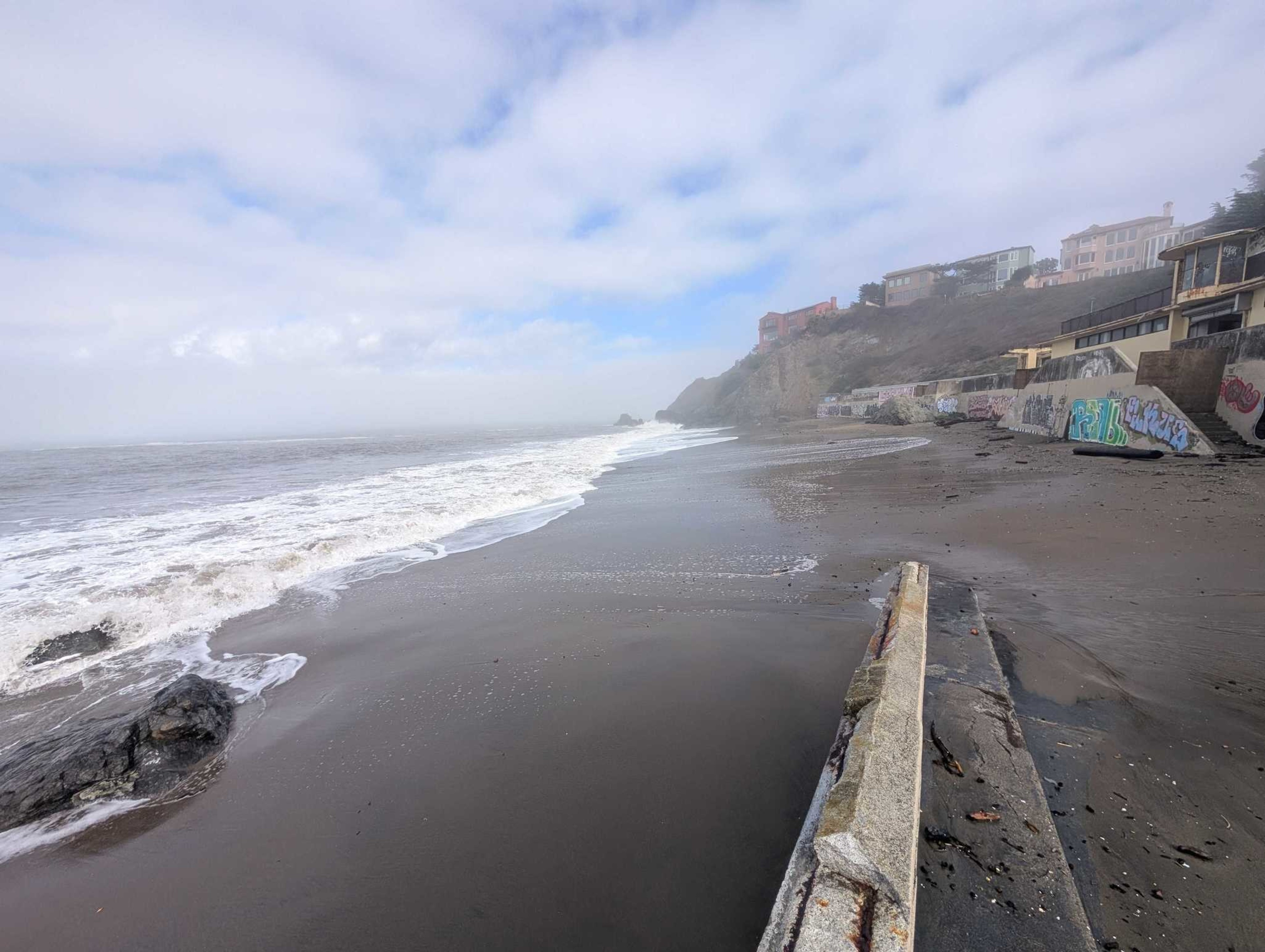 A foggy beach with waves washing ashore, rocky cliffs in the distance, and a seawall covered in graffiti lining the sandy shore.