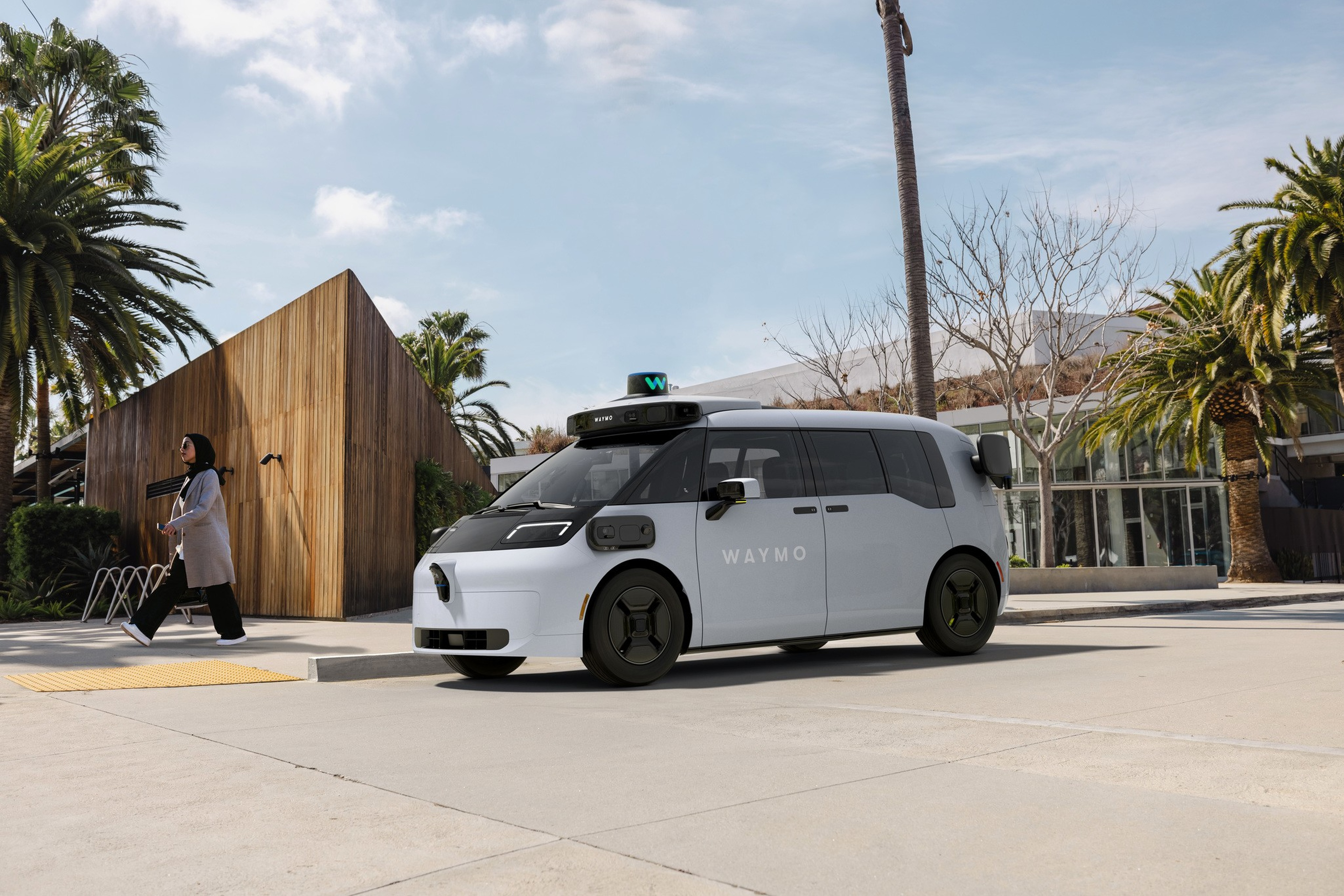 A compact white and black Waymo autonomous vehicle is parked near palm trees and a modern wooden building, while a woman walks crossing the street.