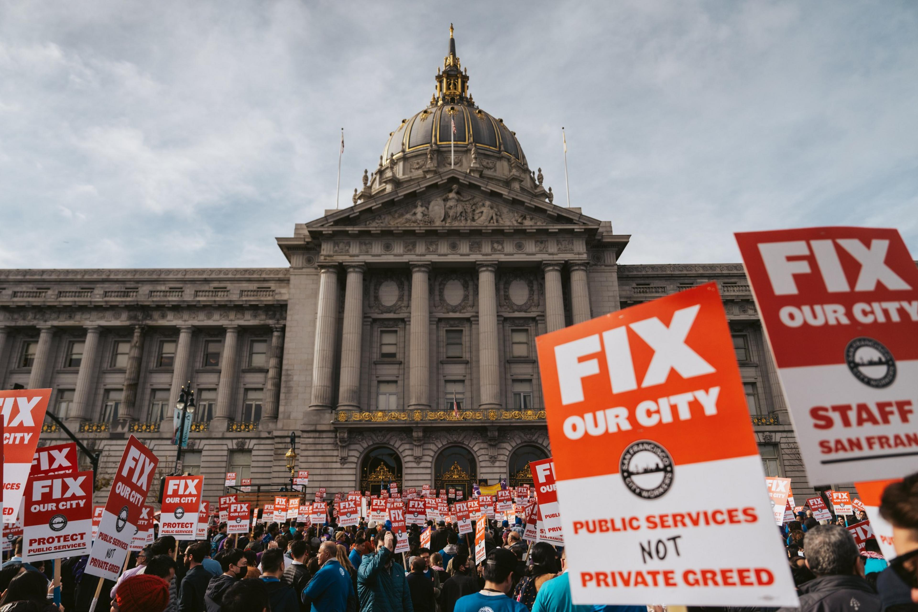 A protest in front of a stately building, with people holding &quot;FIX OUR CITY&quot; signs.