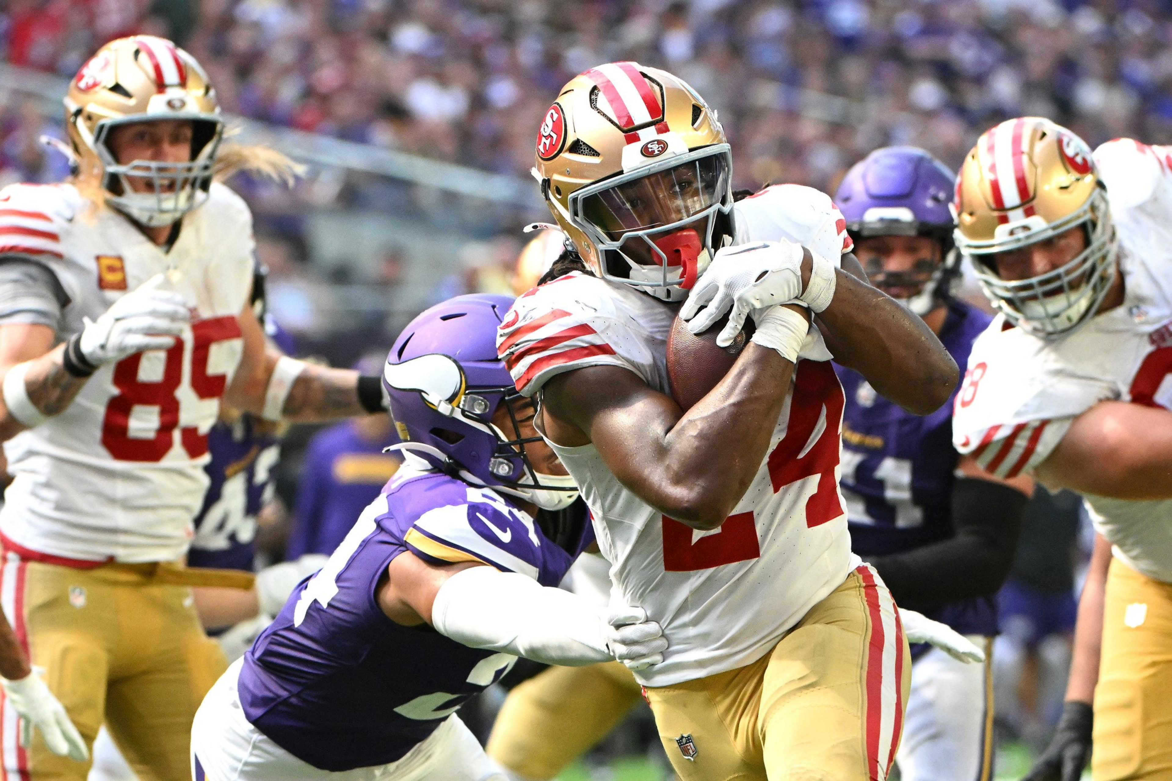 A football player in a white jersey and golden helmet runs with the ball as a player in a purple jersey attempts to tackle him, with teammates nearby.