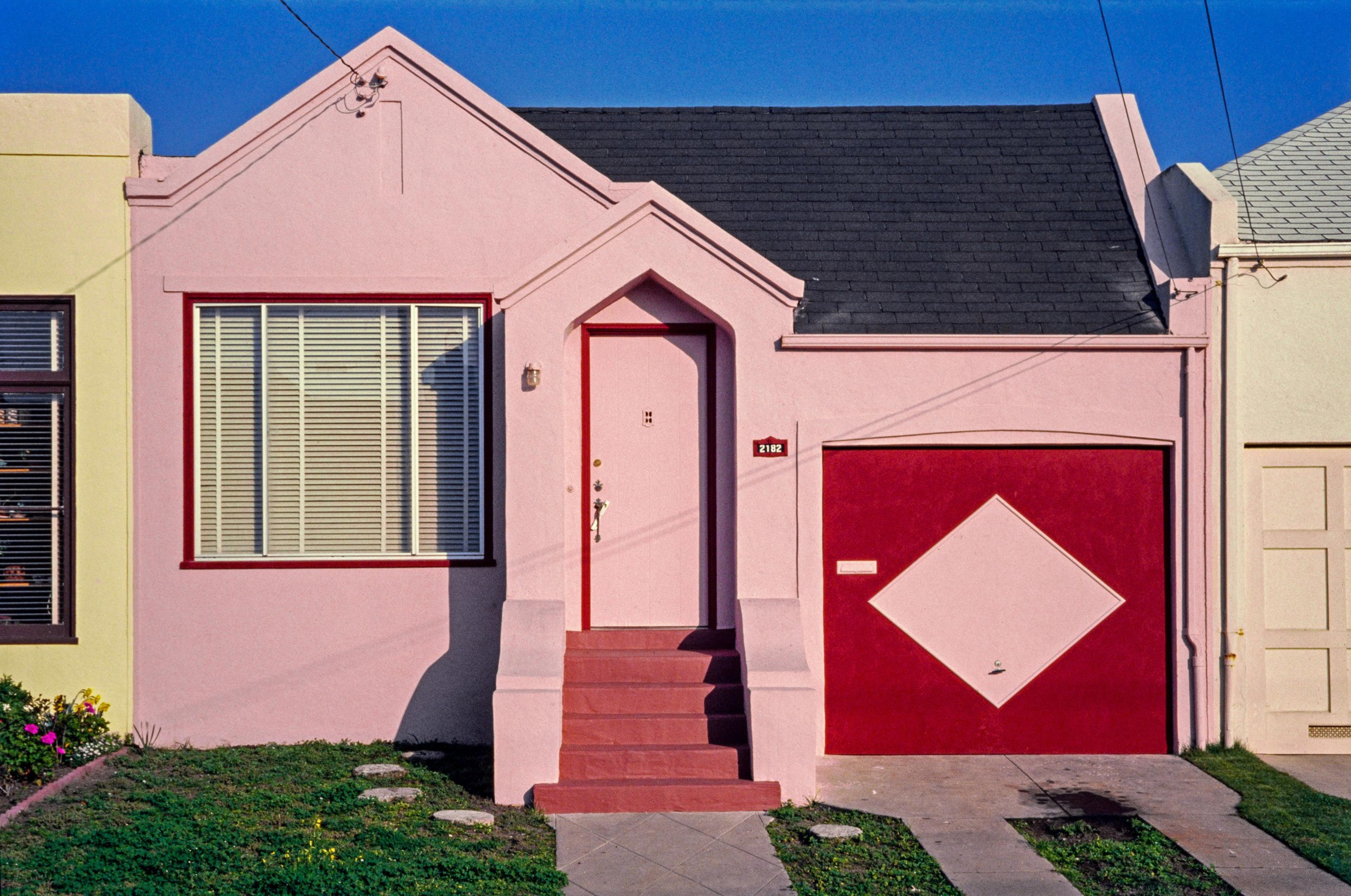 A small pink house with a red door and a garage featuring a diamond pattern. It has a neat lawn with stepping stones and a small flowerbed.