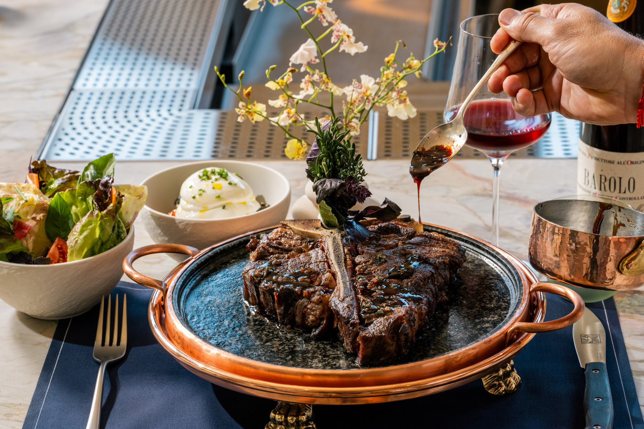 A large grilled T-bone steak on a decorative plate is being drizzled with sauce, accompanied by a salad, burrata cheese, and a glass of red wine.