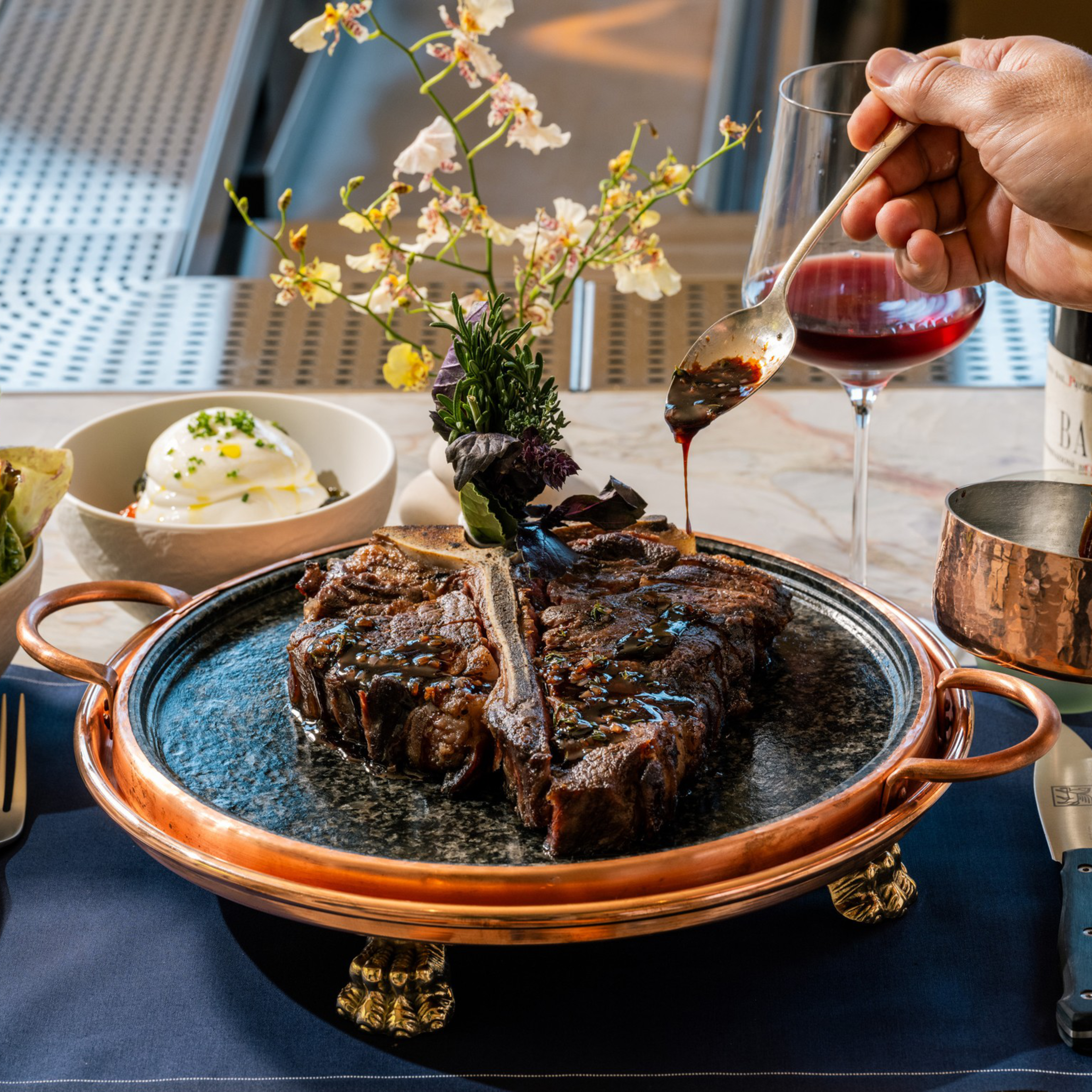 A large grilled T-bone steak on a decorative plate is being drizzled with sauce, accompanied by a salad, burrata cheese, and a glass of red wine.