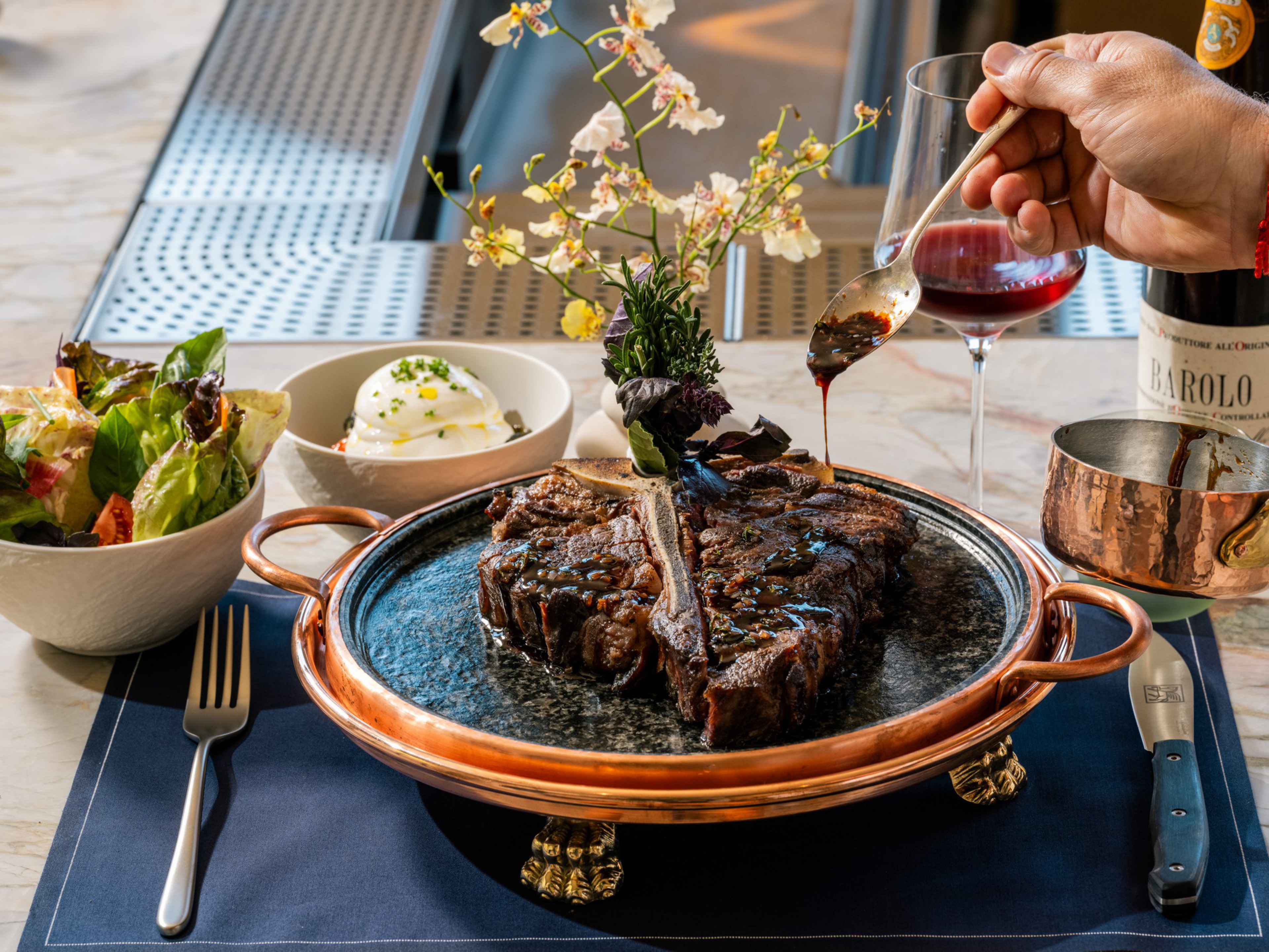 A large grilled T-bone steak on a decorative plate is being drizzled with sauce, accompanied by a salad, burrata cheese, and a glass of red wine.