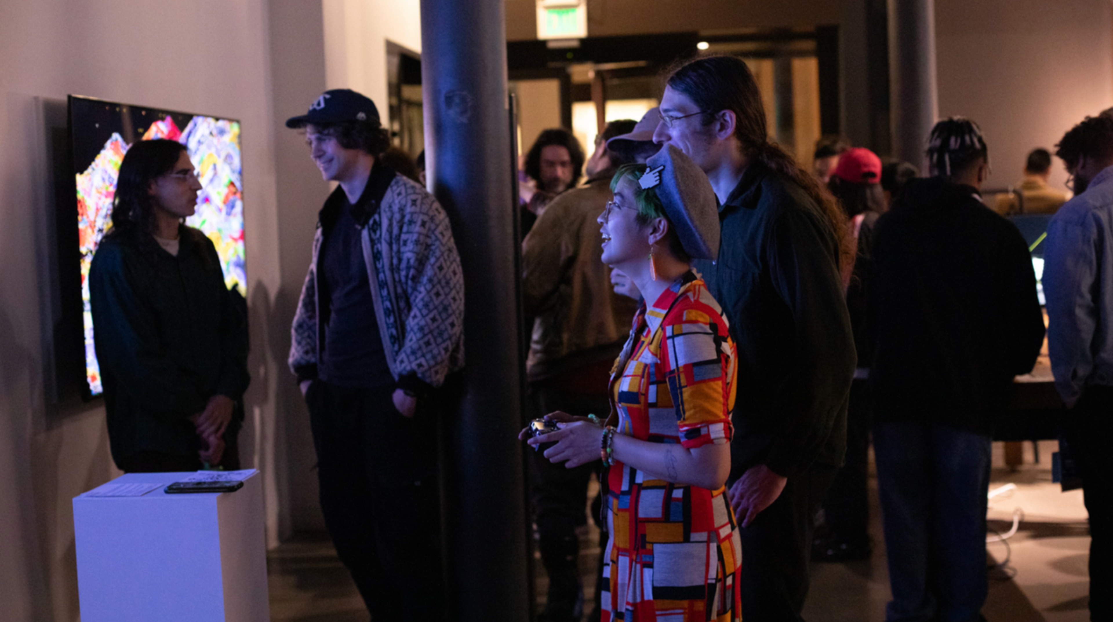 People interact and view digital screens in a dimly lit room, with some holding controllers and others engaged in conversation near a colorful display.