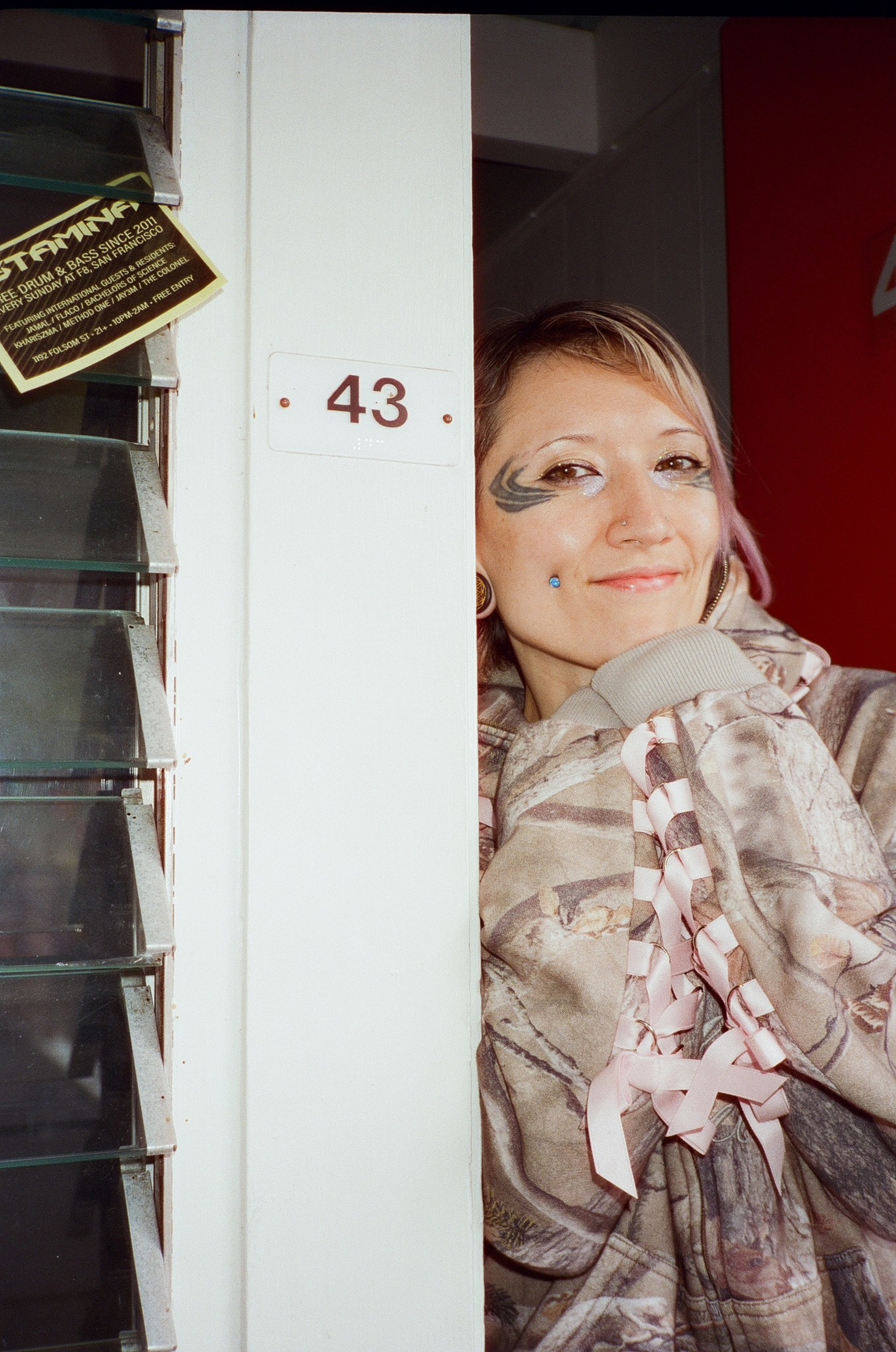 A person with face tattoos and piercings smiles while wearing a camo hoodie with pink ribbons, standing next to a white pillar labeled "43."