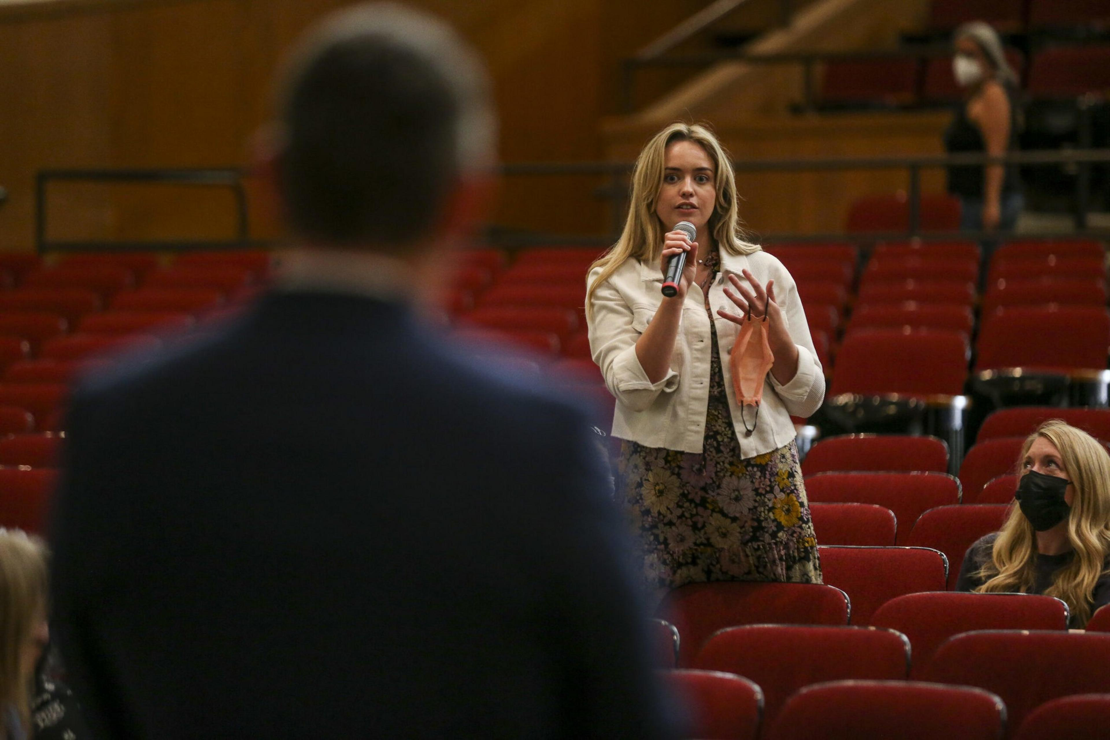 A woman stands among red auditorium seats, holding a microphone and speaking, while a man in the foreground listens and another woman wearing a mask sits nearby.