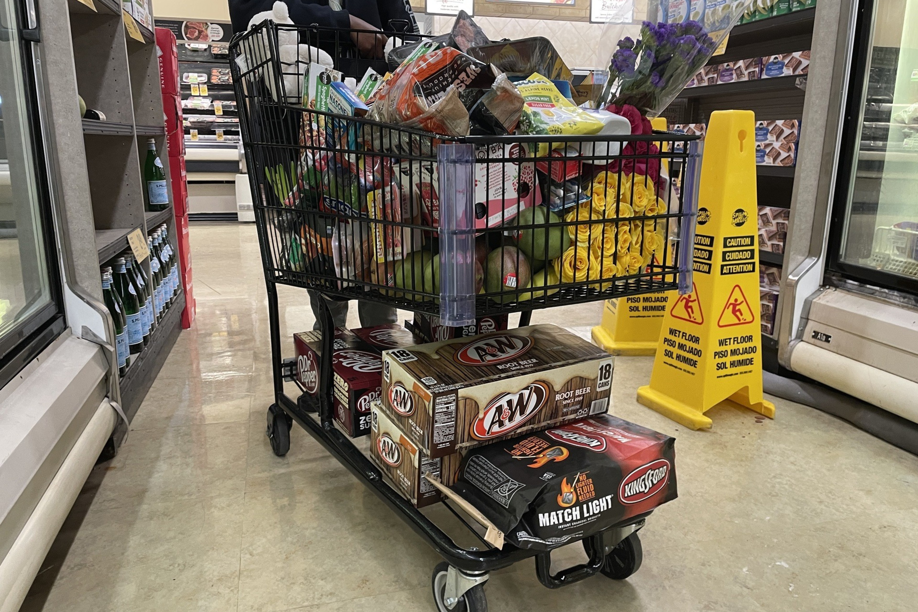 A shopping cart is filled to the brim with groceries, including flowers. Two boxes of root beer and charcoal are beneath the cart. Caution signs are nearby.
