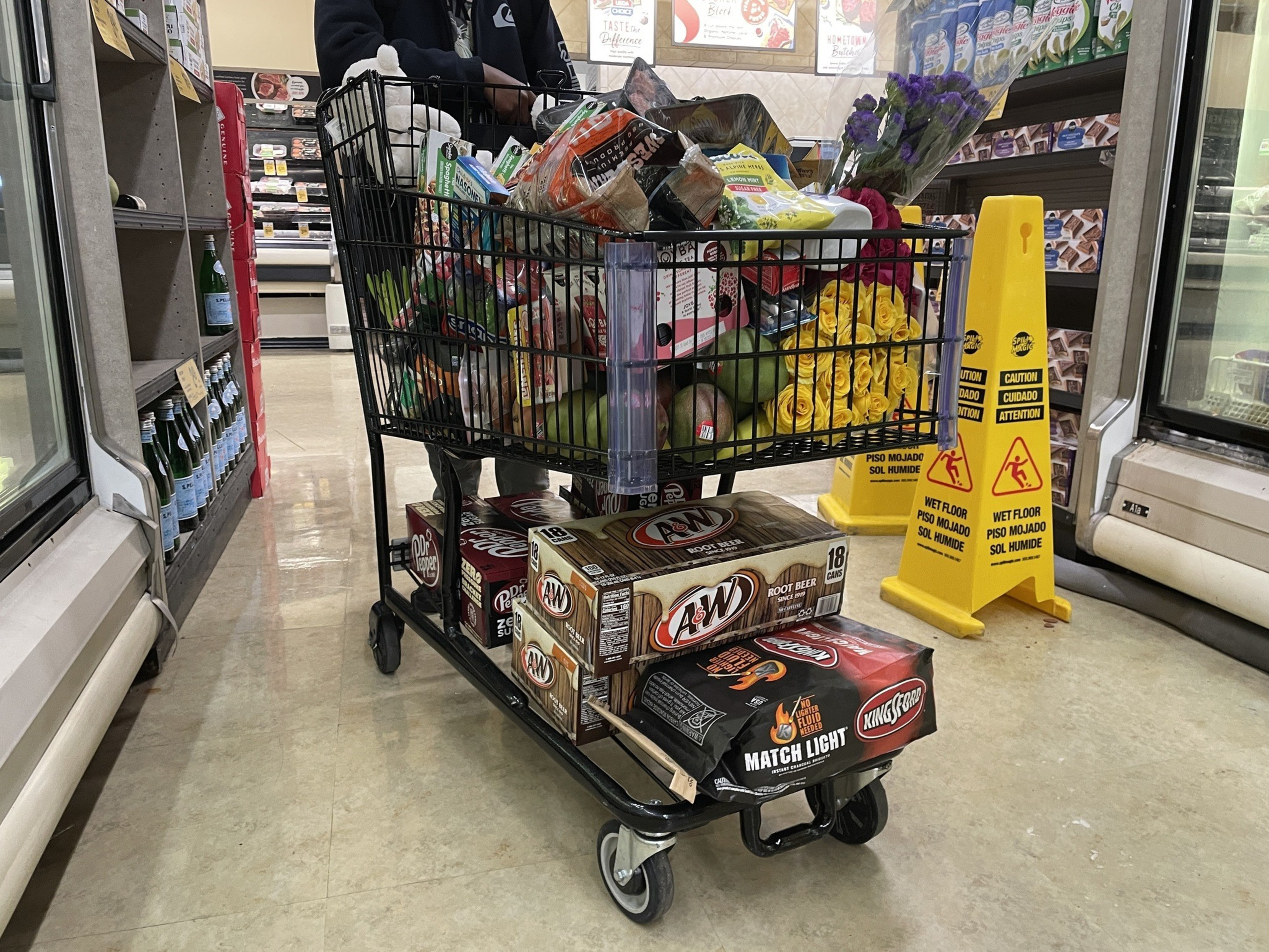 A shopping cart is filled to the brim with groceries, including flowers. Two boxes of root beer and charcoal are beneath the cart. Caution signs are nearby.