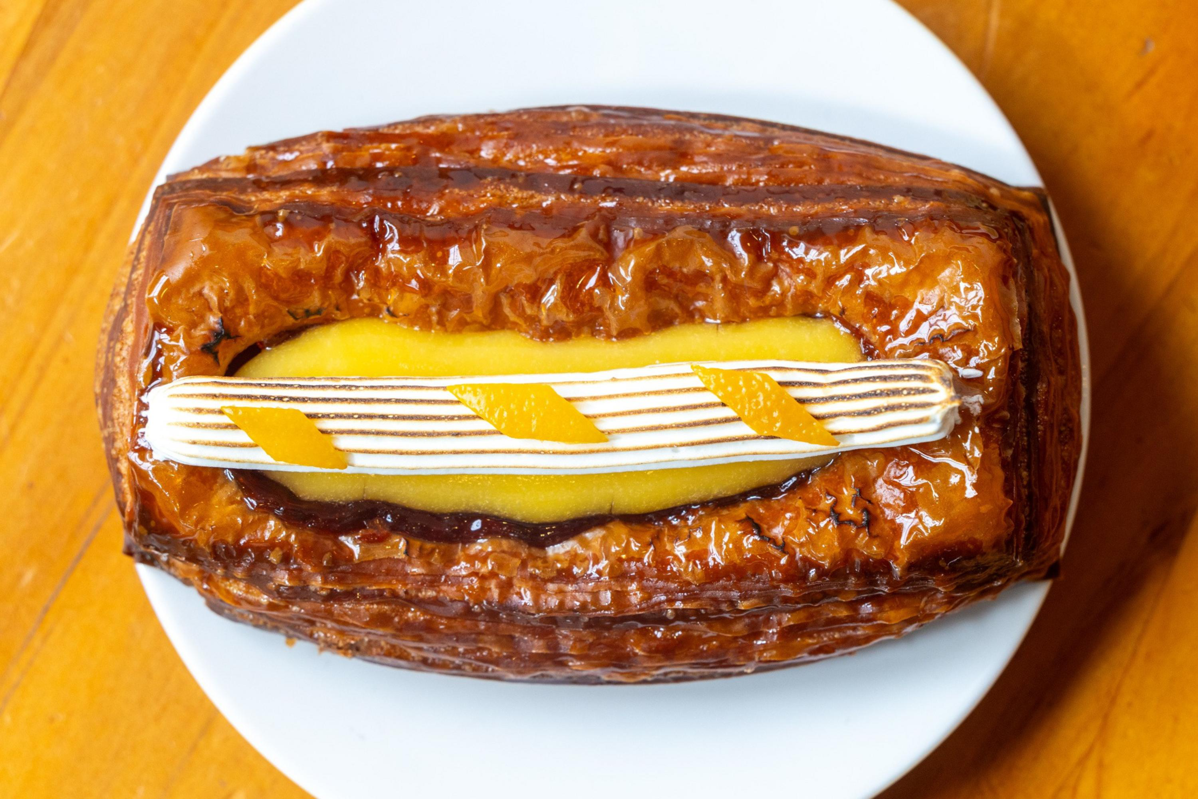 A rectangular glazed pastry with a yellow custard center topped with a white-striped cream and small orange peel garnishes, on a white plate.
