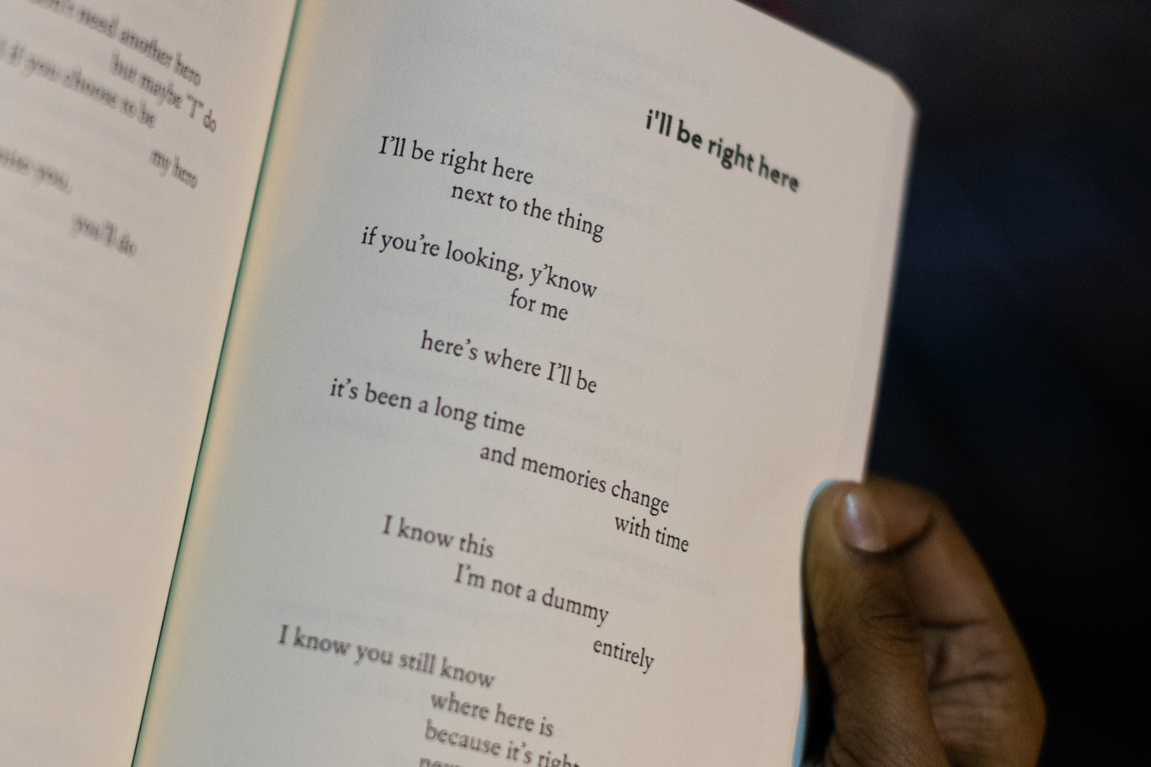 A hand holds an open book displaying a poem titled "i'll be right here" with lines arranged in a staggered, irregular pattern on the page.