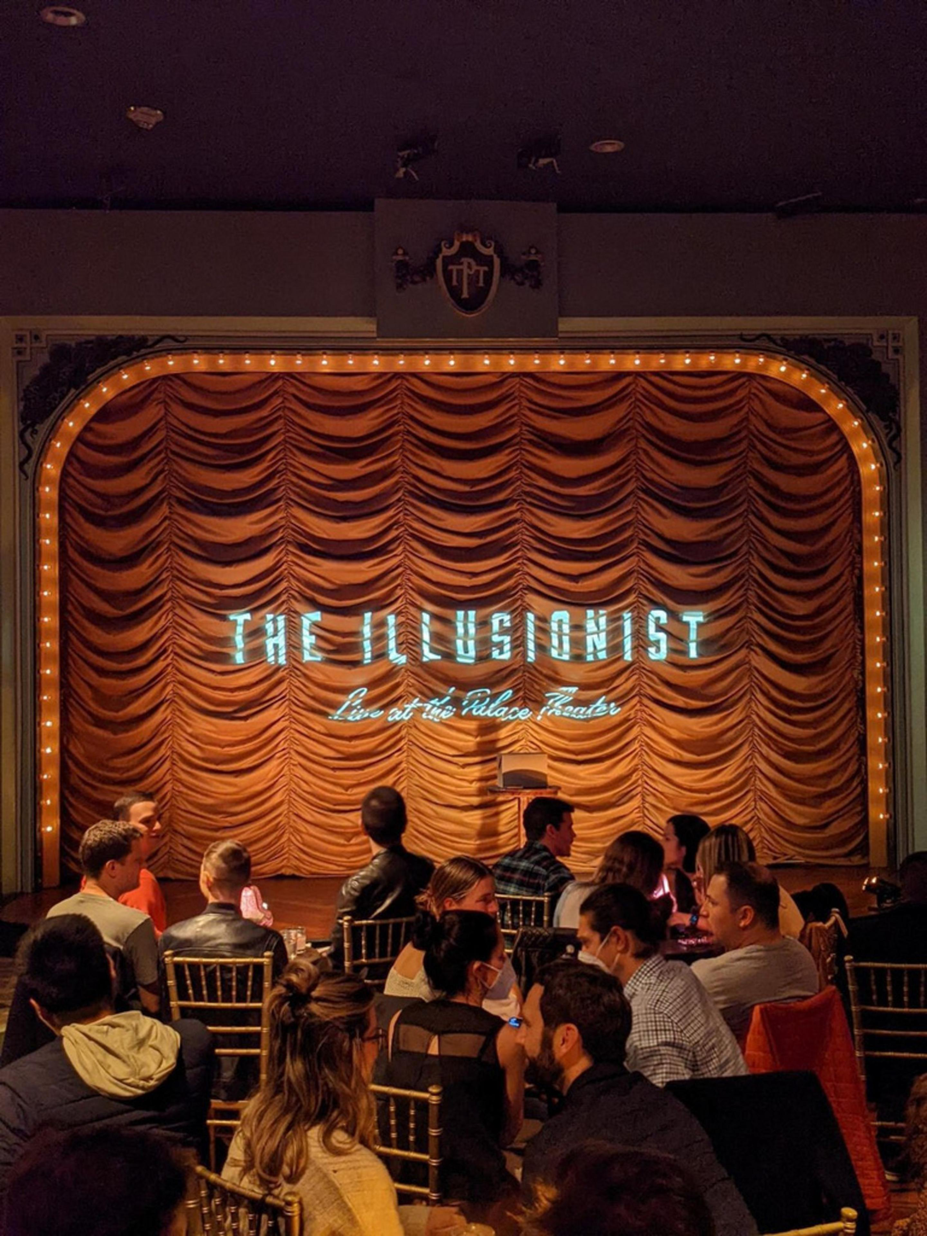 People sit facing a stage with a closed, orange curtain lit by a sign that reads "The Illusionist Live at the Palace Theater."
