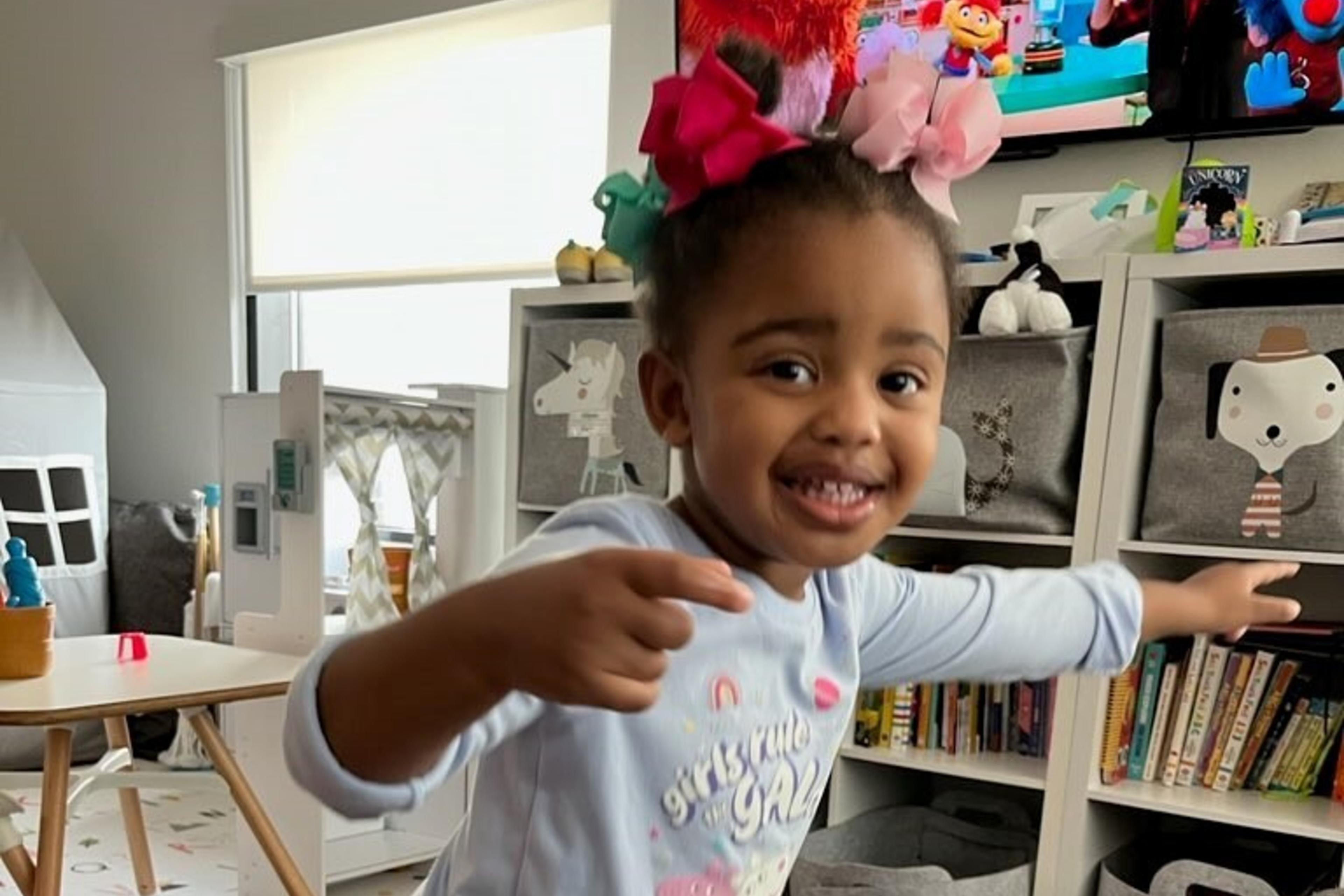 A smiling child with bows in her hair points playfully in a tidy playroom with toy bins, a TV showing a children's program, and colorful foam mats.