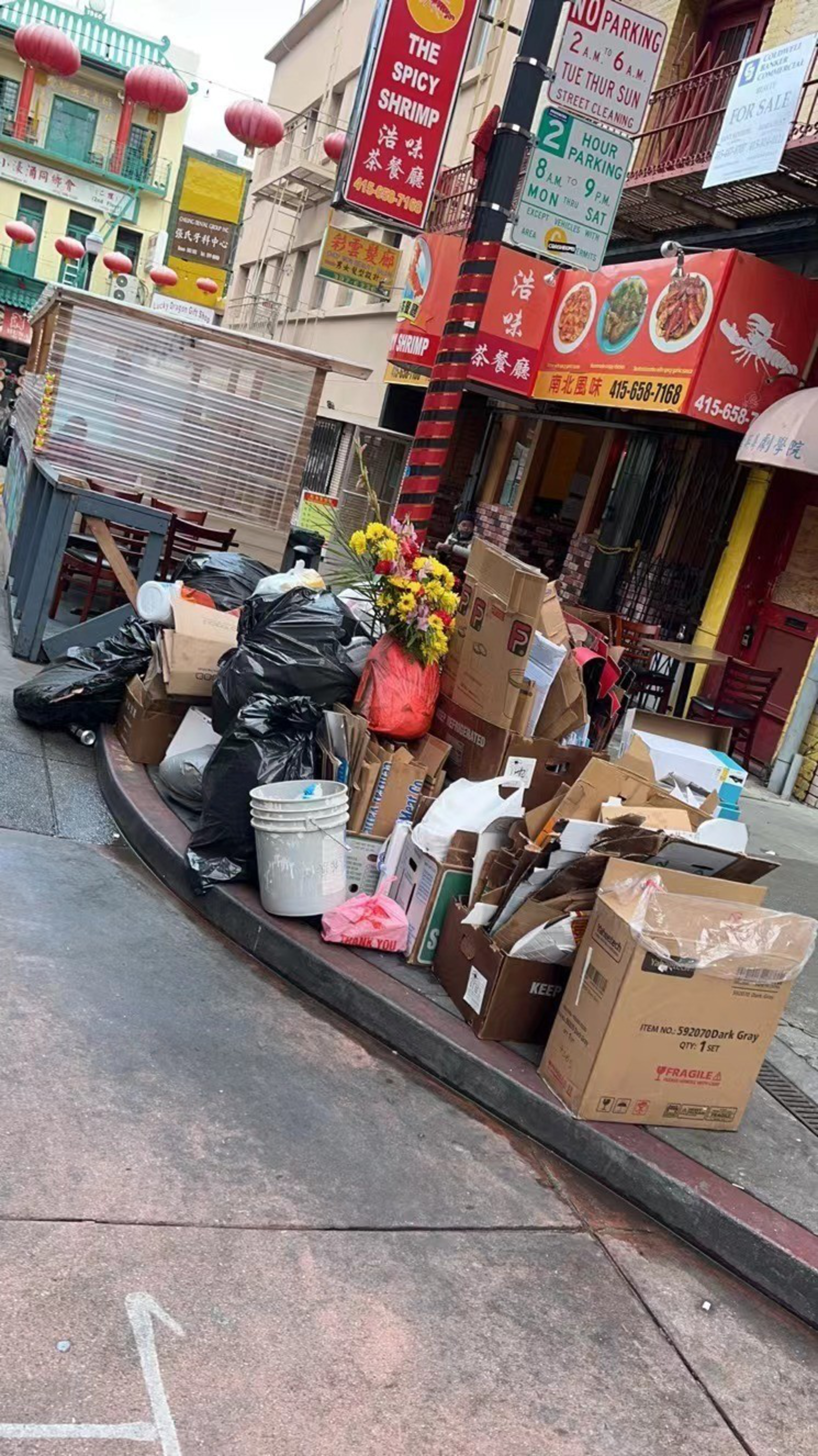Piles of black trash bags, cardboard boxes, and a white bucket with flowers sit on a sidewalk curb in front of a restaurant with red signage and Chinese characters.