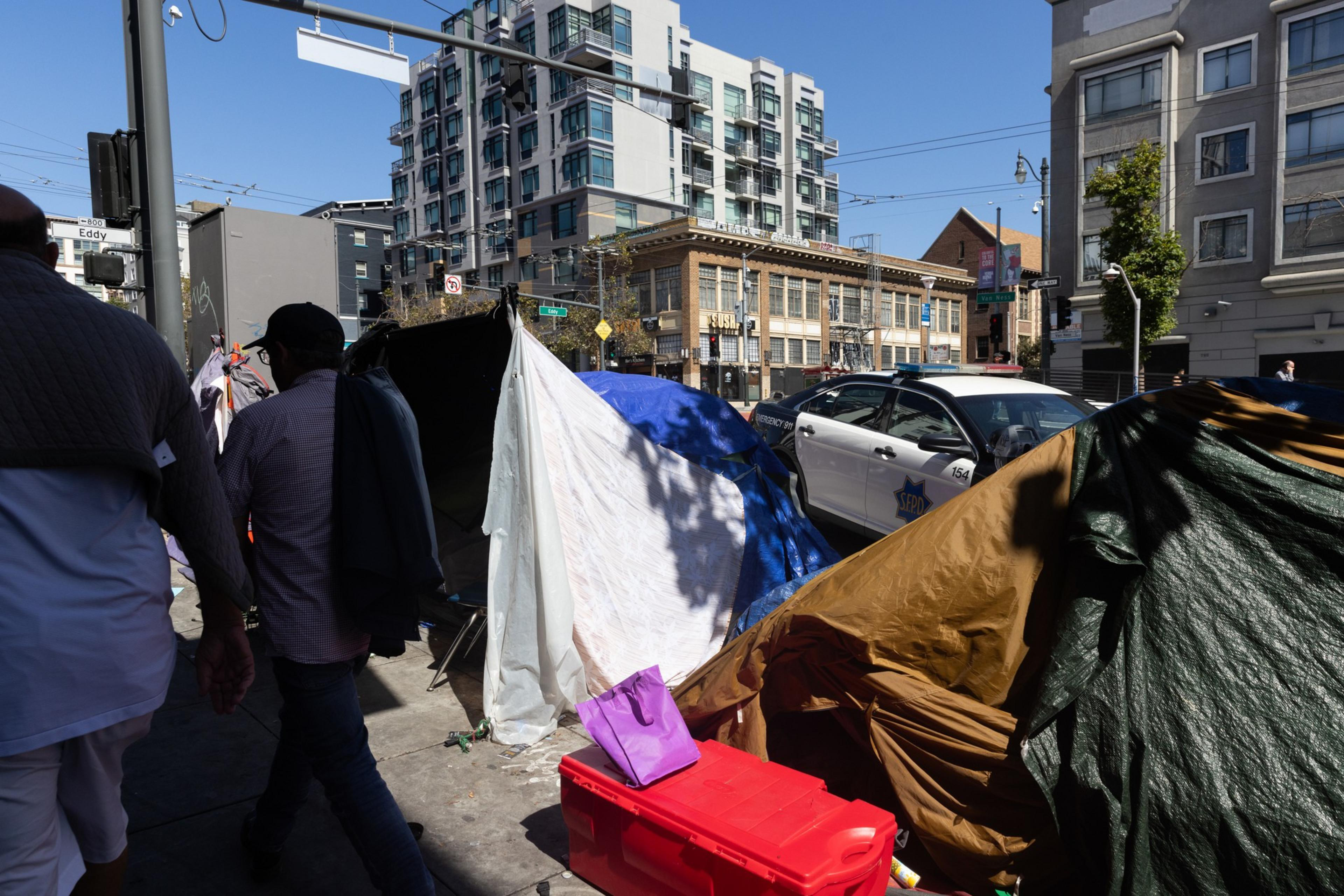 Tents covered with tarps line a city sidewalk near a police car, with people walking past tall buildings under a clear blue sky.