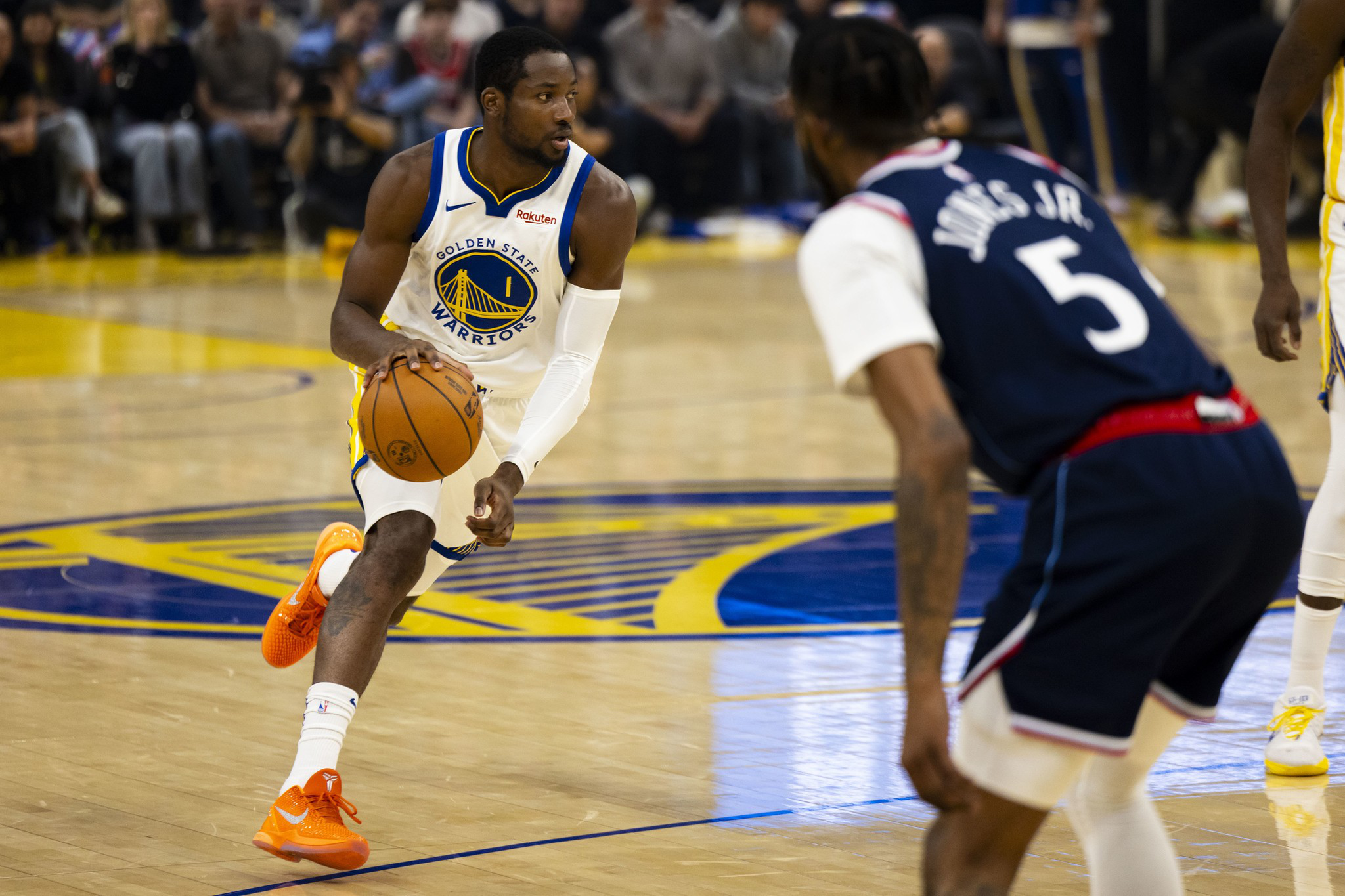 A Golden State Warriors player dribbles the basketball while being closely guarded by an opponent wearing a navy blue jersey with the number 5.