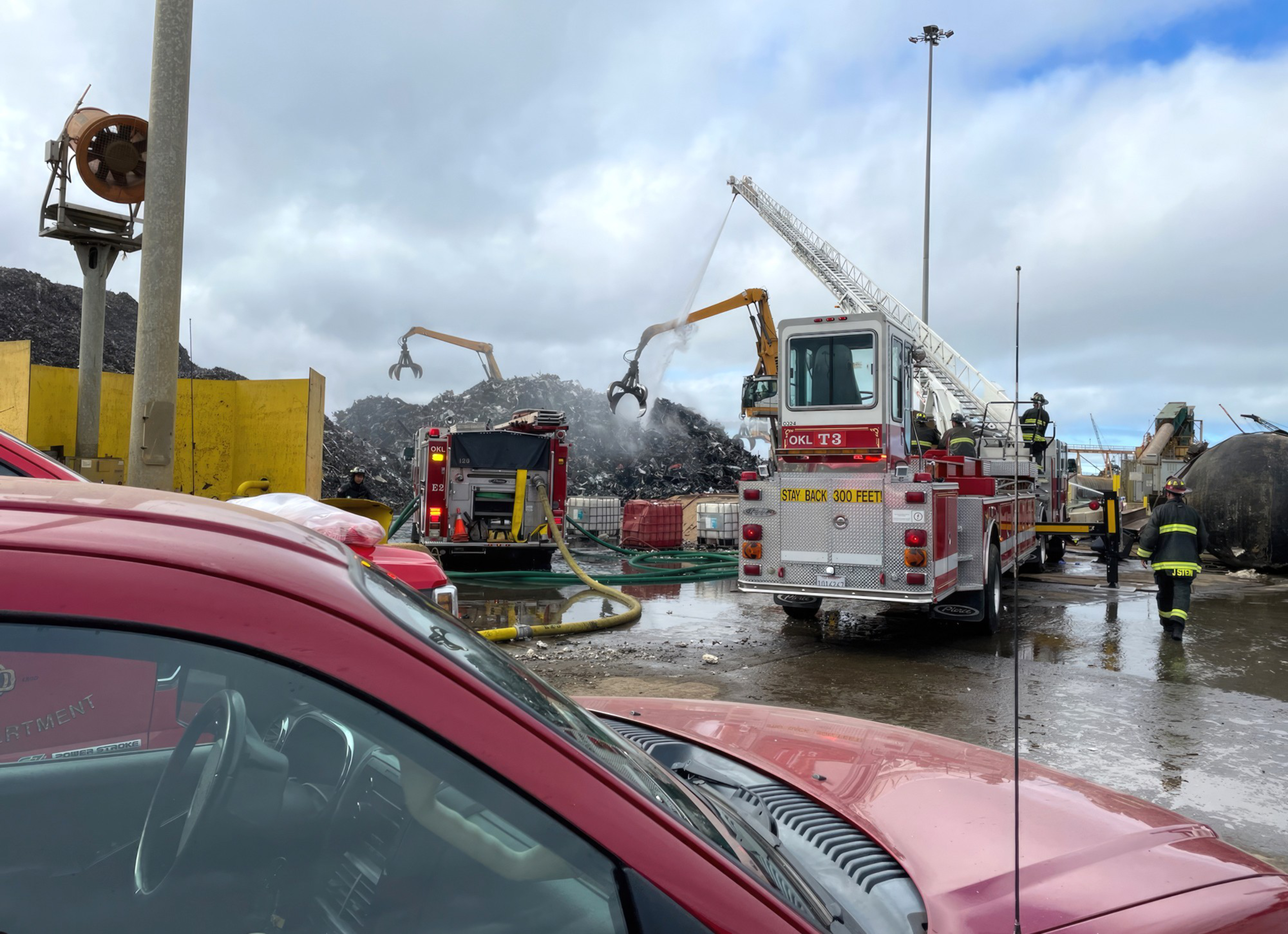 Firefighters use a ladder truck to spray water on a large pile of burning debris at a landfill while heavy machinery moves materials.