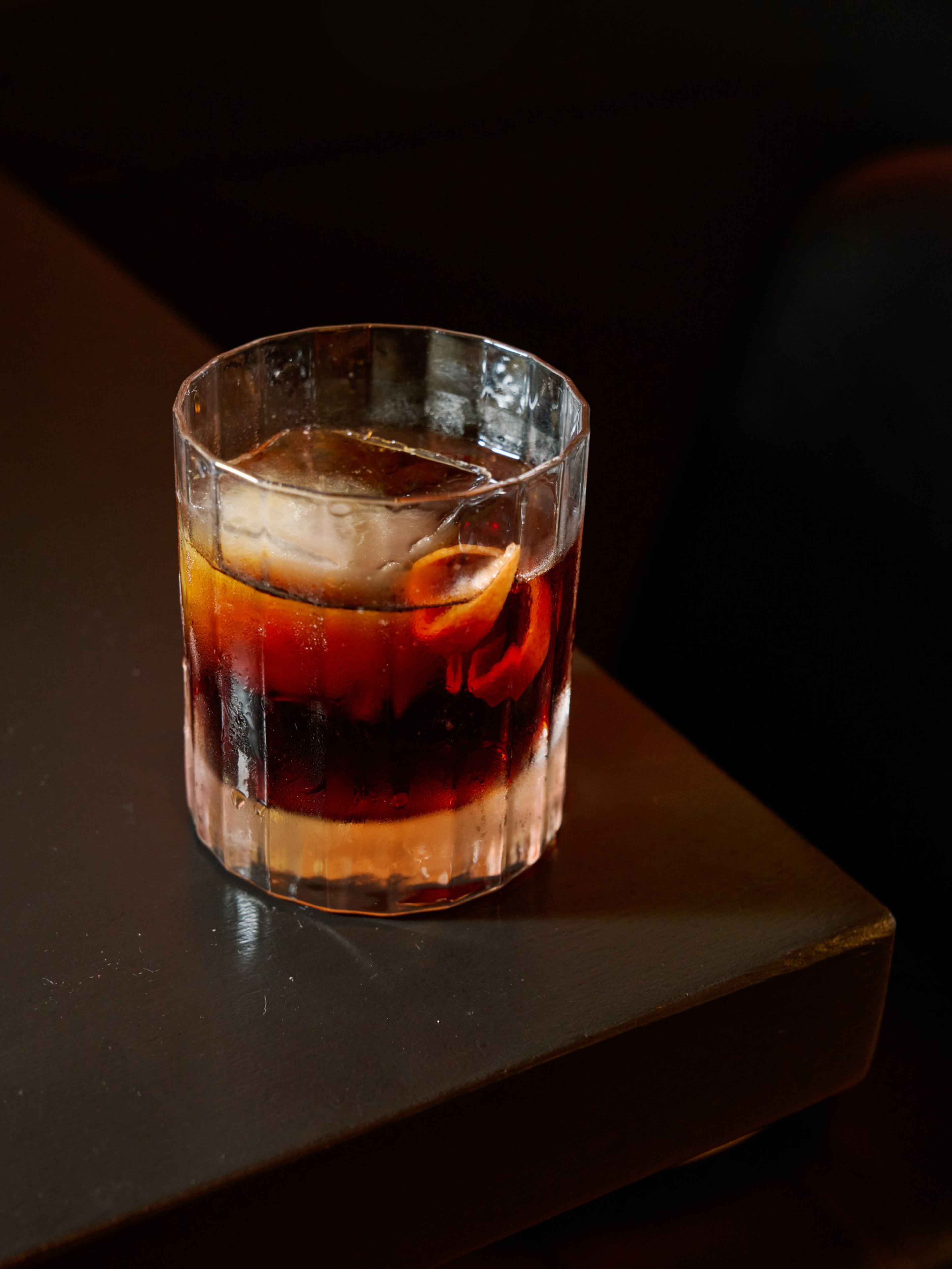 A short, faceted glass contains a dark drink with a large ice cube and a twisted orange peel on a dark surface.