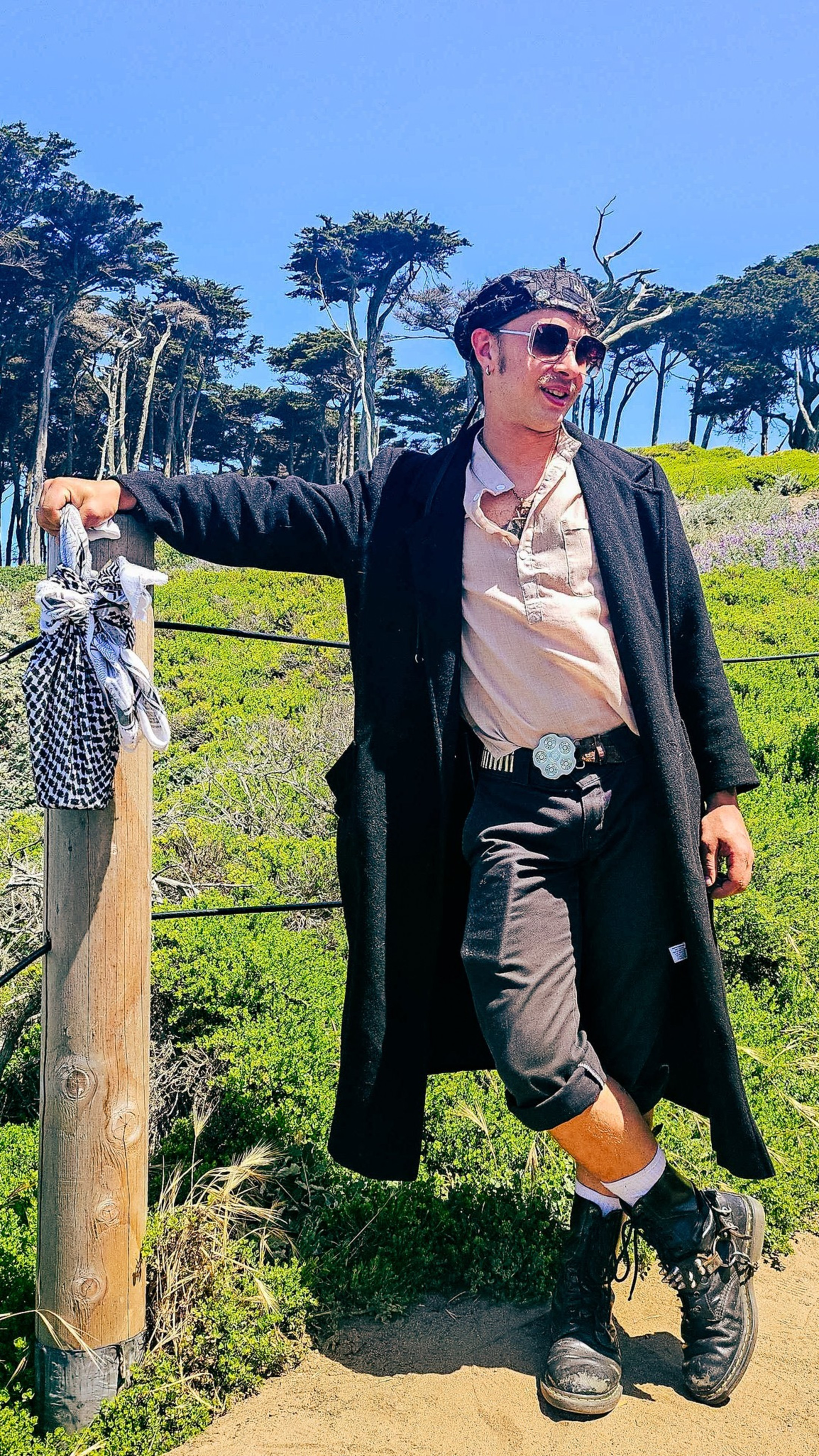 A person in sunglasses and a long black coat leans on a post with a bandana tied to it. They are outdoors with vibrant green foliage and tall trees.